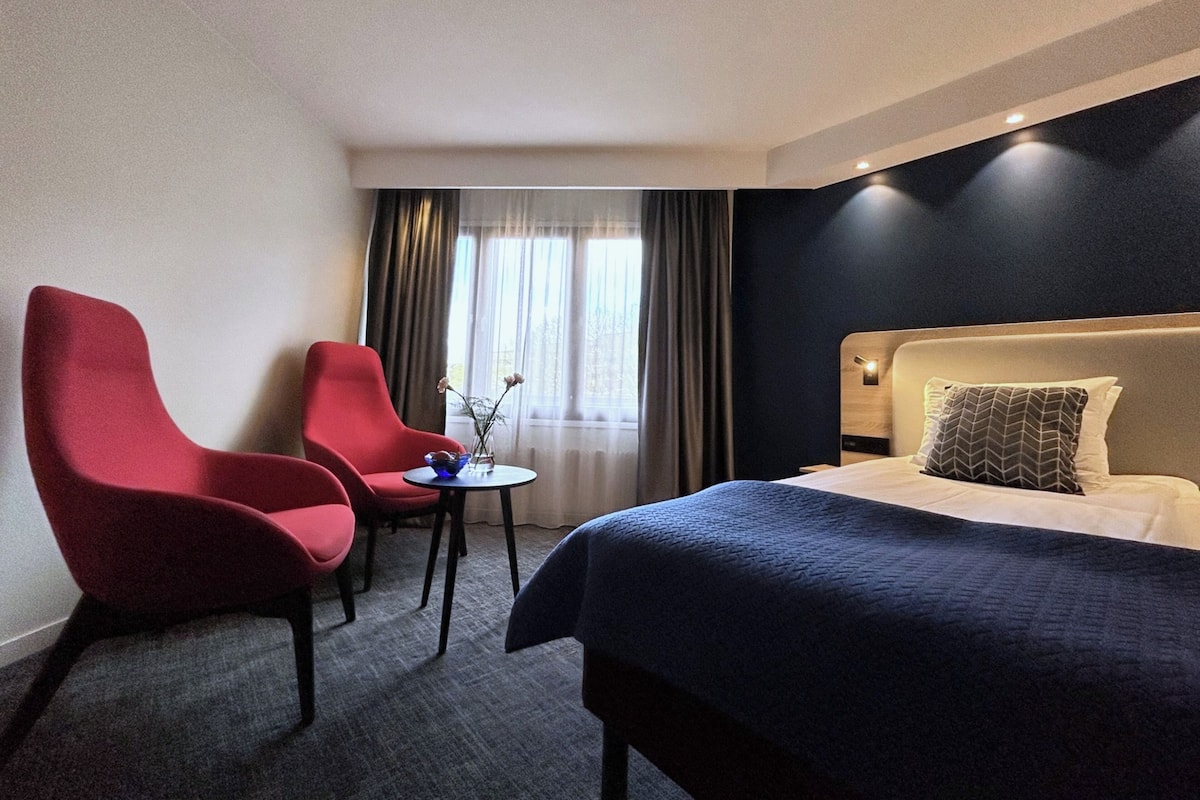 A hotel room with a bed, two red armchairs, a small table, and a window with curtains.