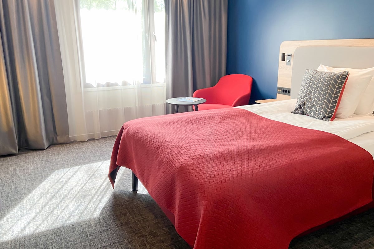 A hotel room with a large bed, a red chair, a small round table, and a view of the outdoors through sheer curtains.