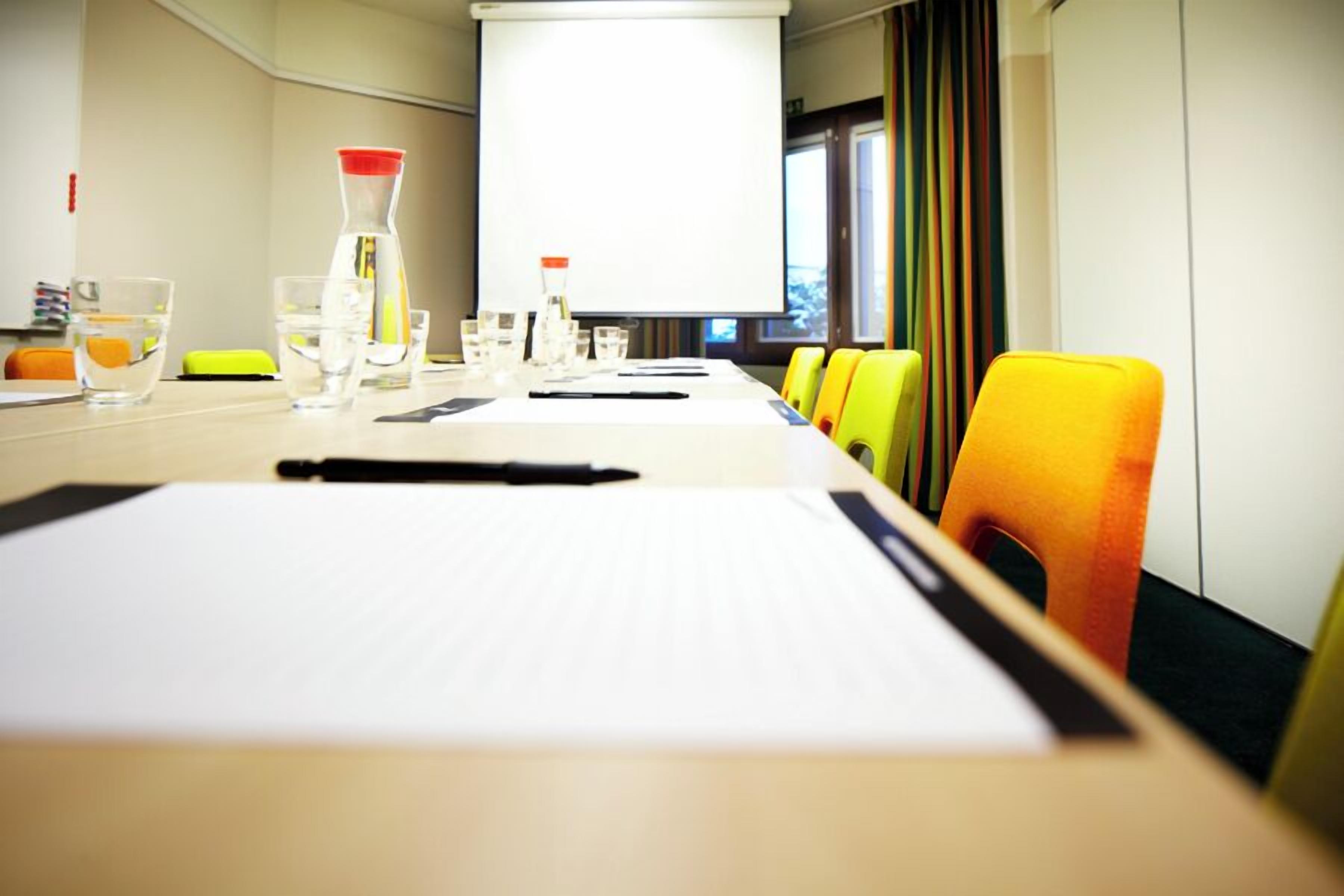 A conference room with a long table, chairs, a projector screen, and a window with curtains.