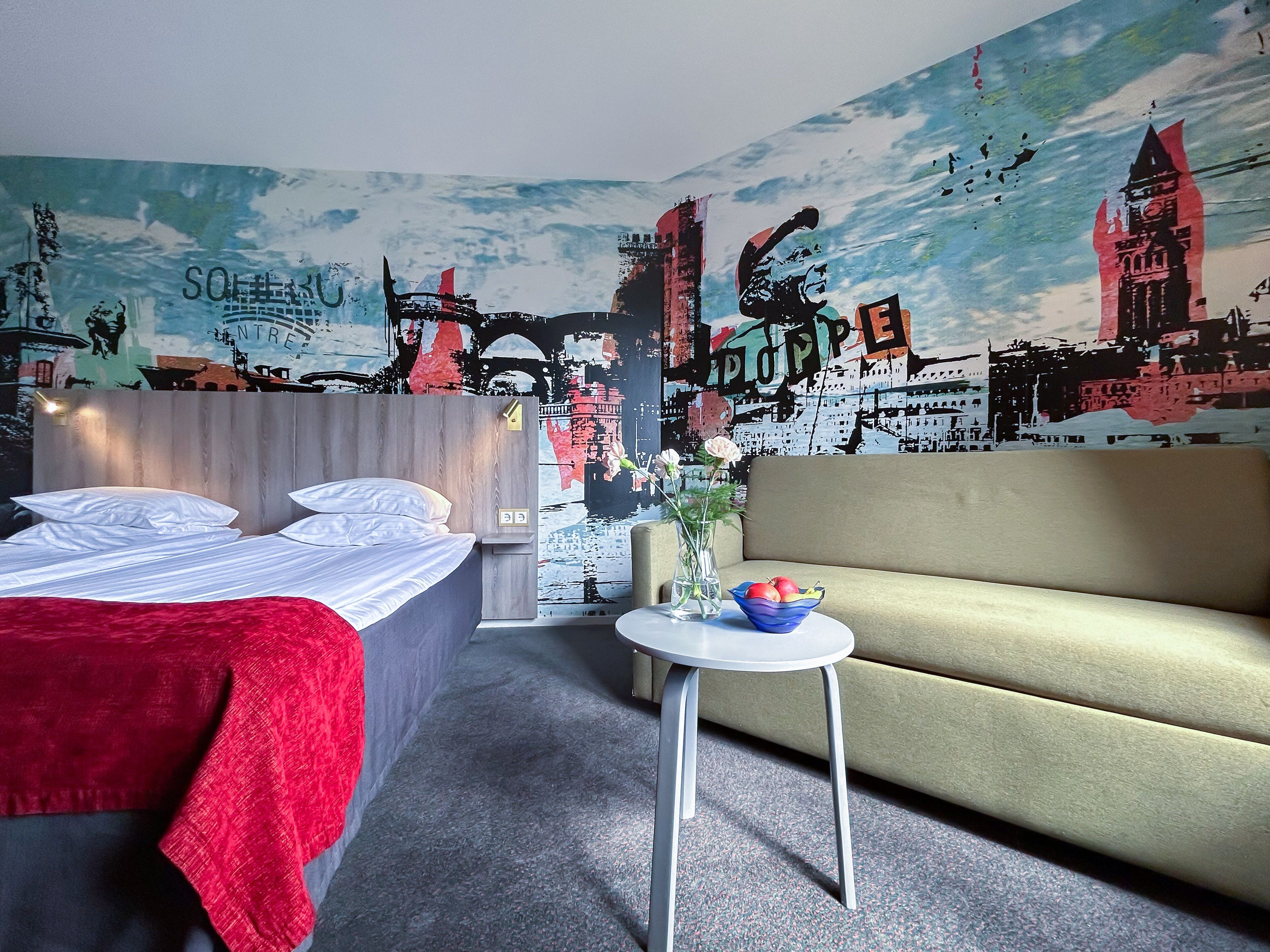 A hotel room with a bed, a sofa, a small table with a vase and fruit bowl, and a mural of a cityscape.