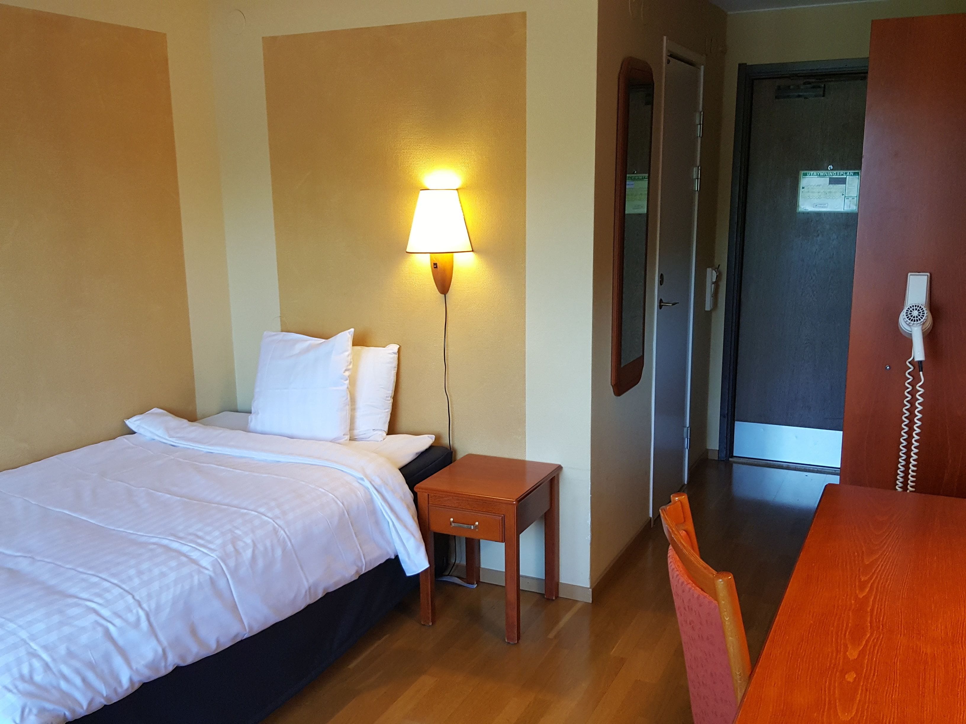 Quarto de hotel com cama, mesa de cabeceira, luminária e uma cadeira de madeira.