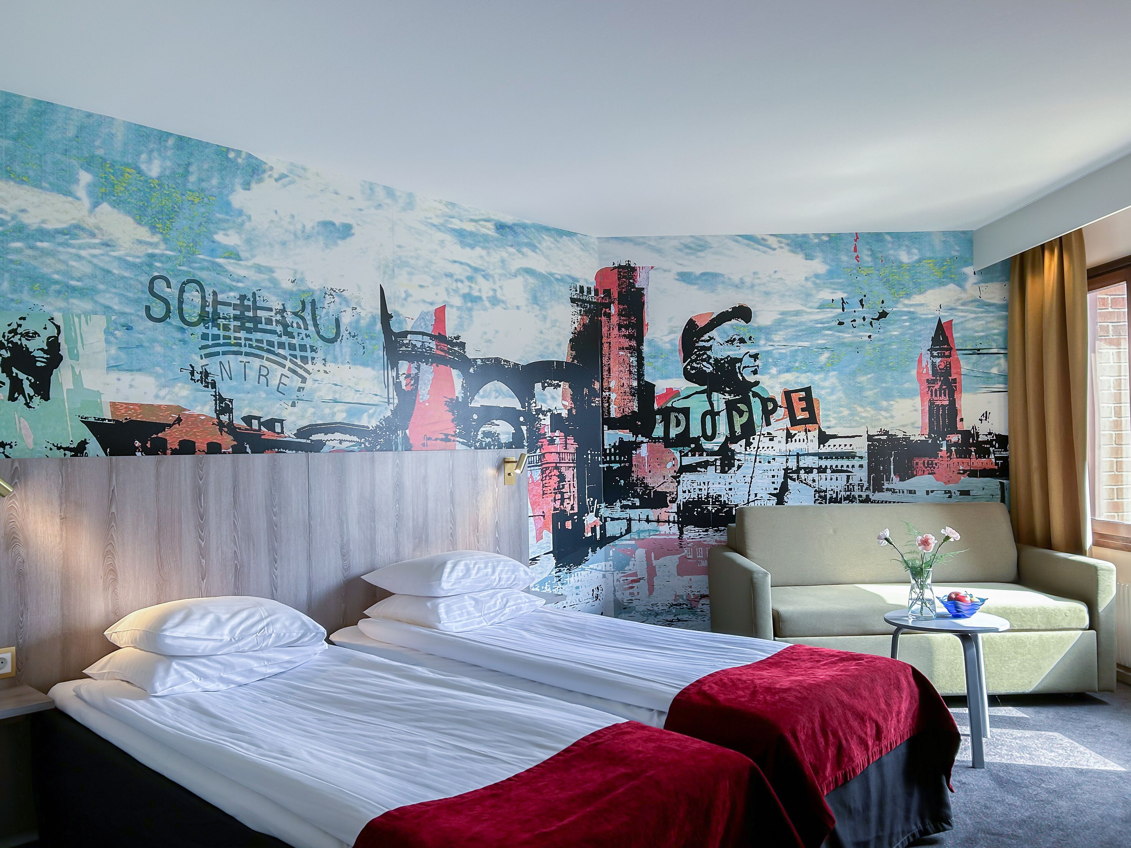 A hotel room with a bed, two pillows, a red blanket, a chair, a small table with a vase and a bowl, and a large cityscape mural on the wall.