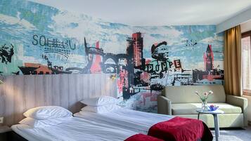 A hotel room with a bed, two pillows, a red blanket, a chair, a small table with a vase and a bowl, and a large cityscape mural on the wall.