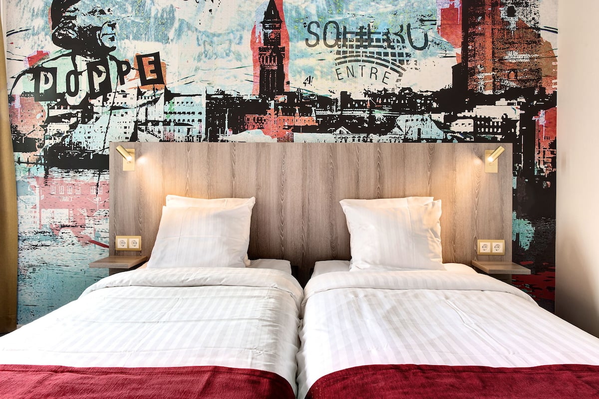 A double bed with white bedding and a red accent, set against a cityscape mural.