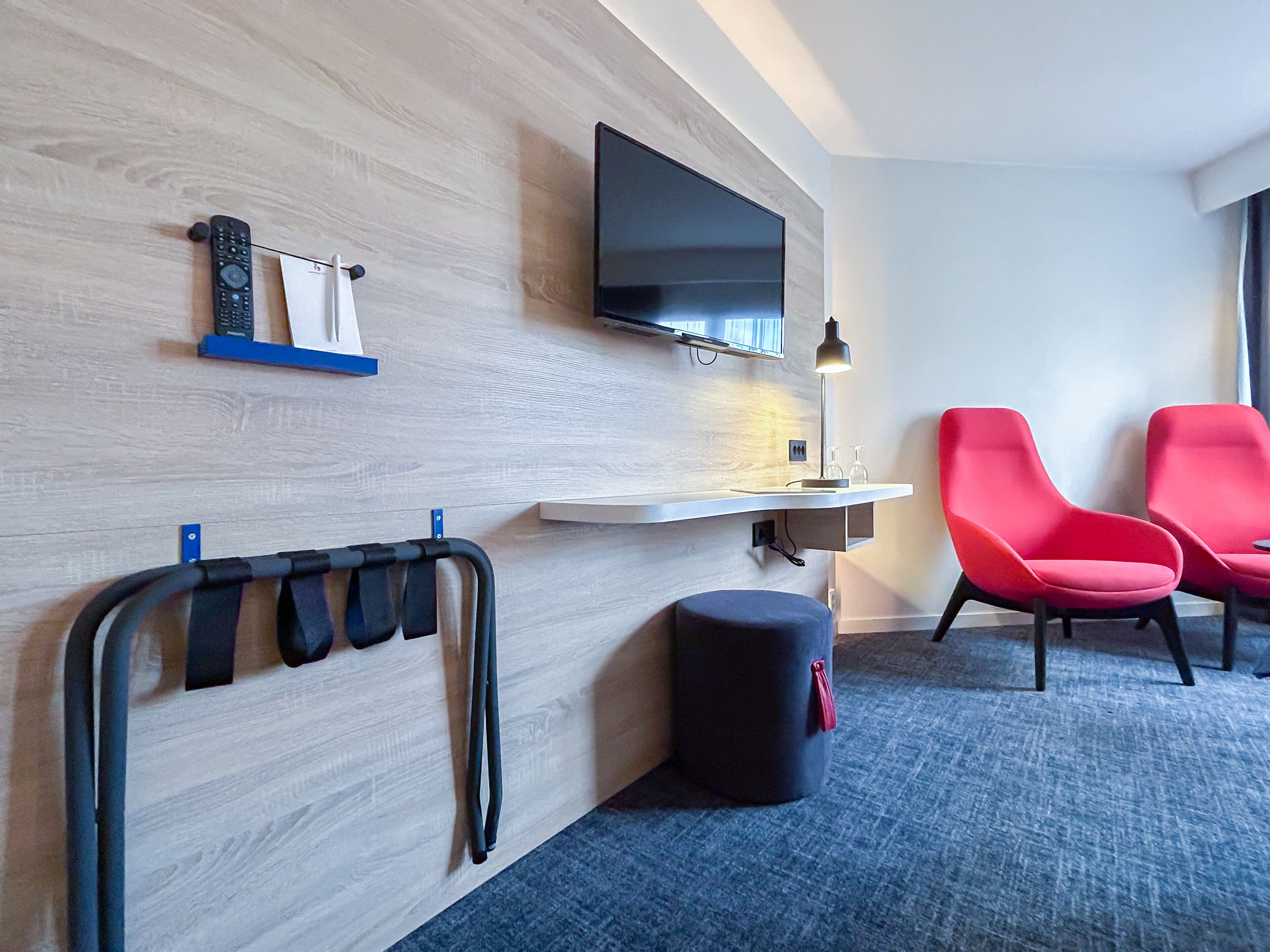 Ein Hotelzimmer mit einem an der Wand befestigten Fernseher, einem Schreibtisch mit Lampe, zwei roten Stühlen, einem schwarzen Hocker und einem Wandregal mit schwarzen Riemen.