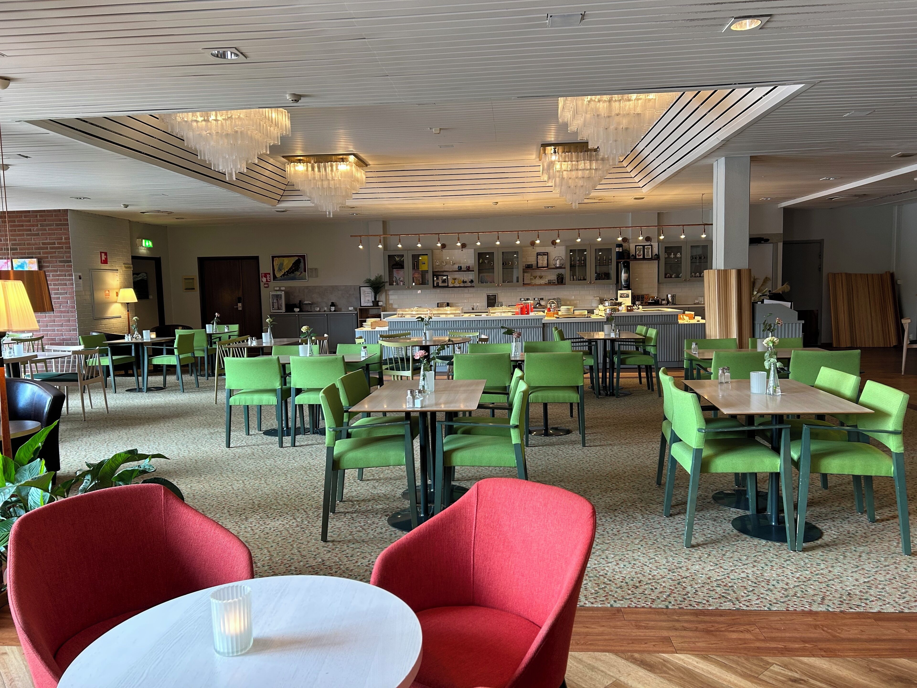 A modern restaurant with green and red chairs, a central bar area, and a ceiling with hanging lights.