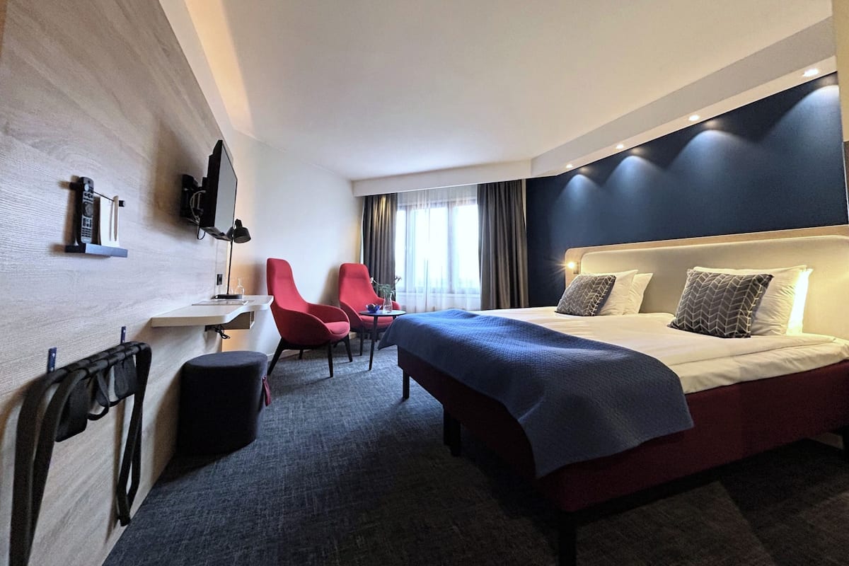 A hotel room with a bed, two red chairs, a desk, and a TV mounted on the wall.