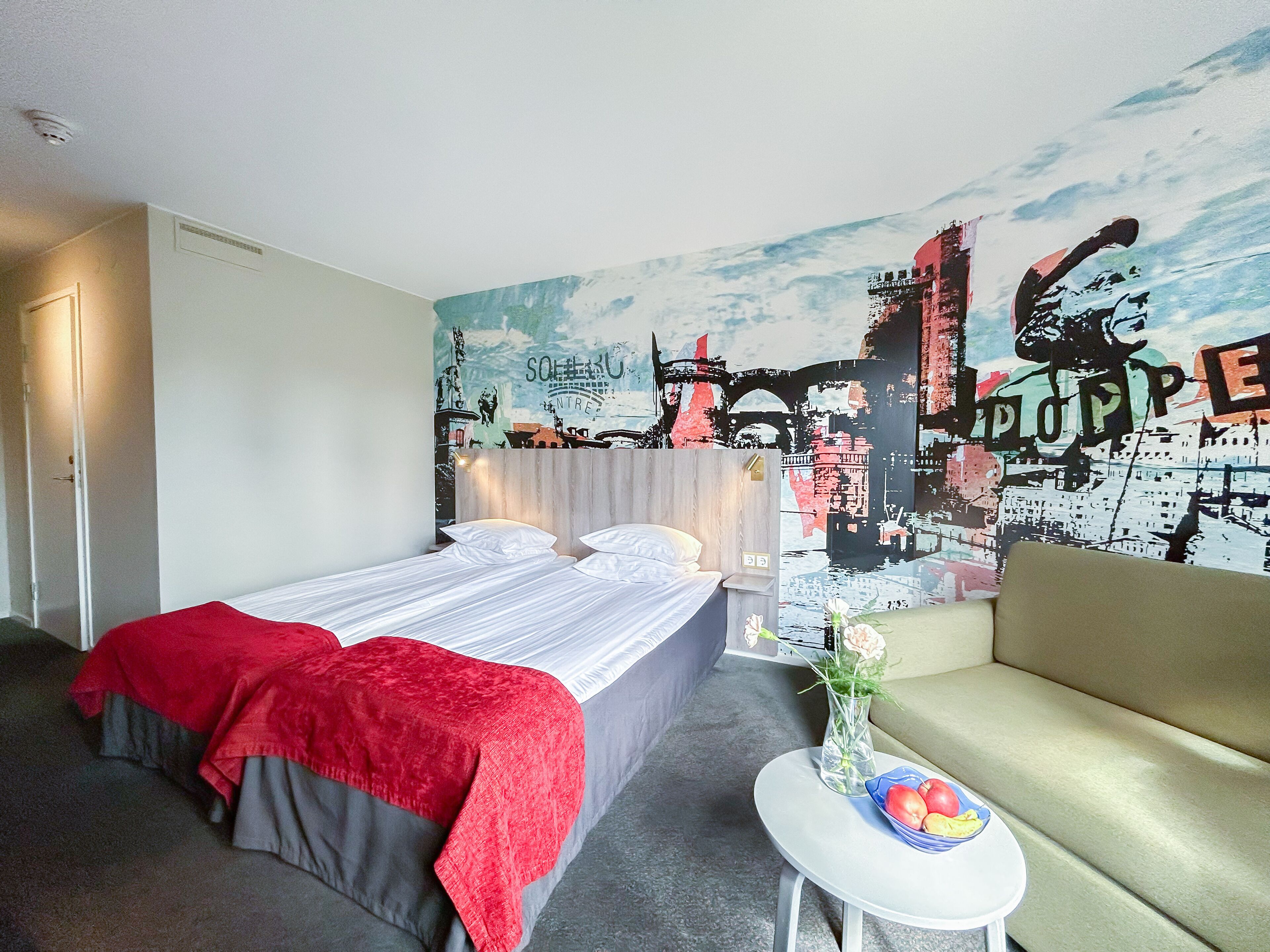 A hotel room with a bed, a sofa, a small table with a vase and fruit, and a mural on the wall.