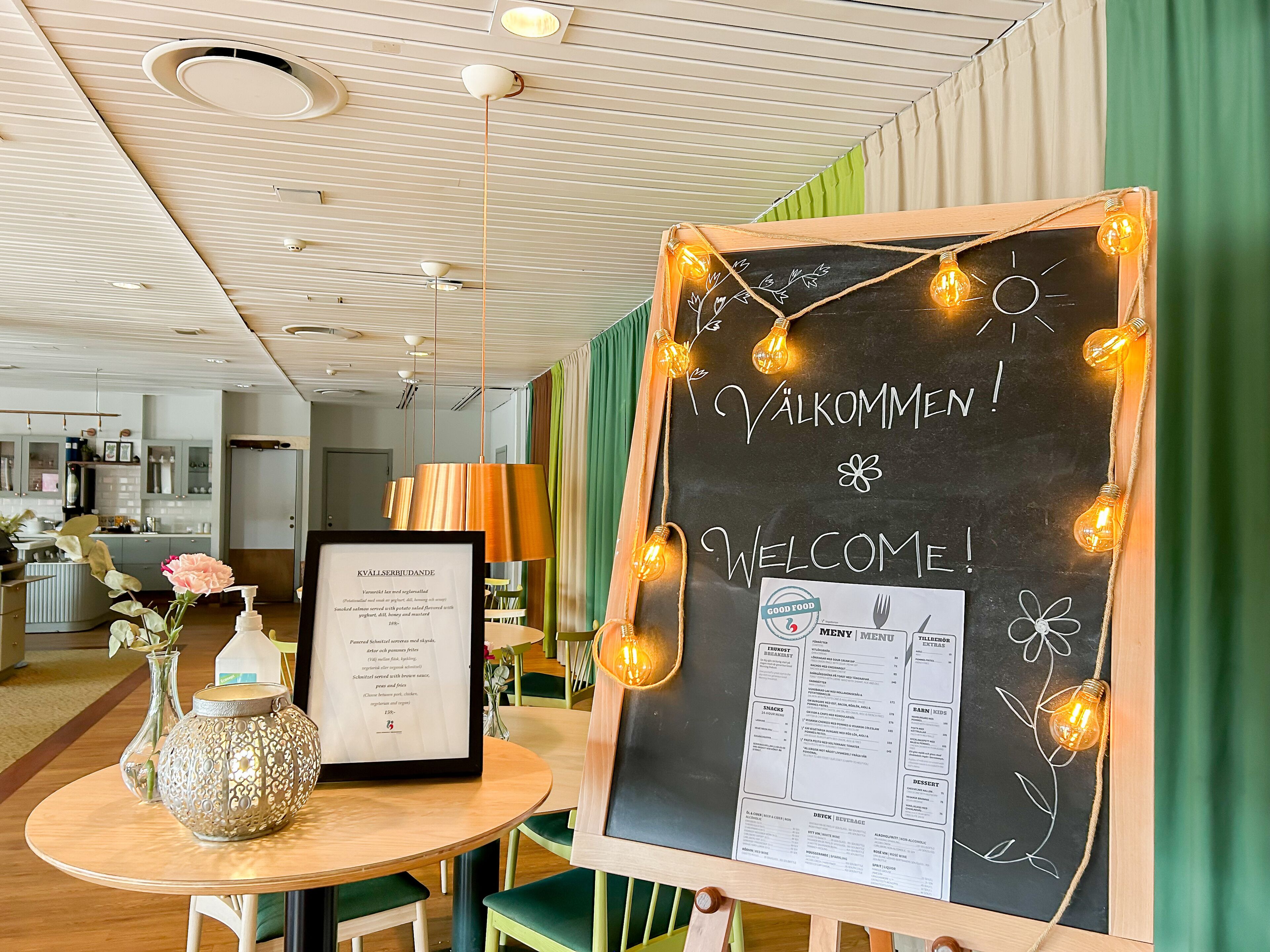 A reception area with a chalkboard displaying 'Welcome!' and a menu, a framed certificate on a table, and a vase with flowers.