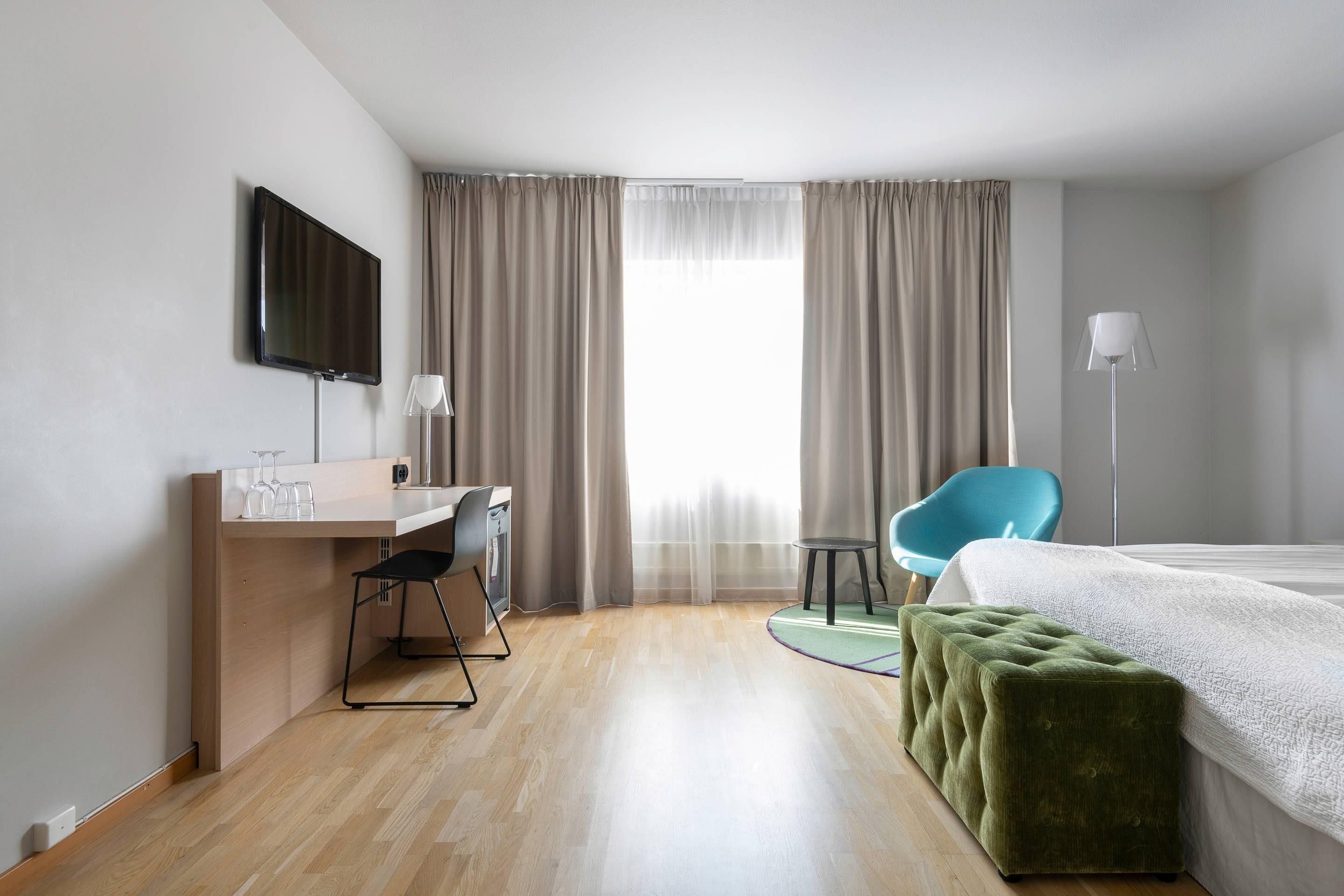 Una habitació d'hotel amb un llit, una taula de nit, una cadira, un televisor i una finestra amb cortines.