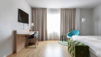 Una habitació d'hotel amb un llit, una taula de nit, una cadira, un televisor i una finestra amb cortines.