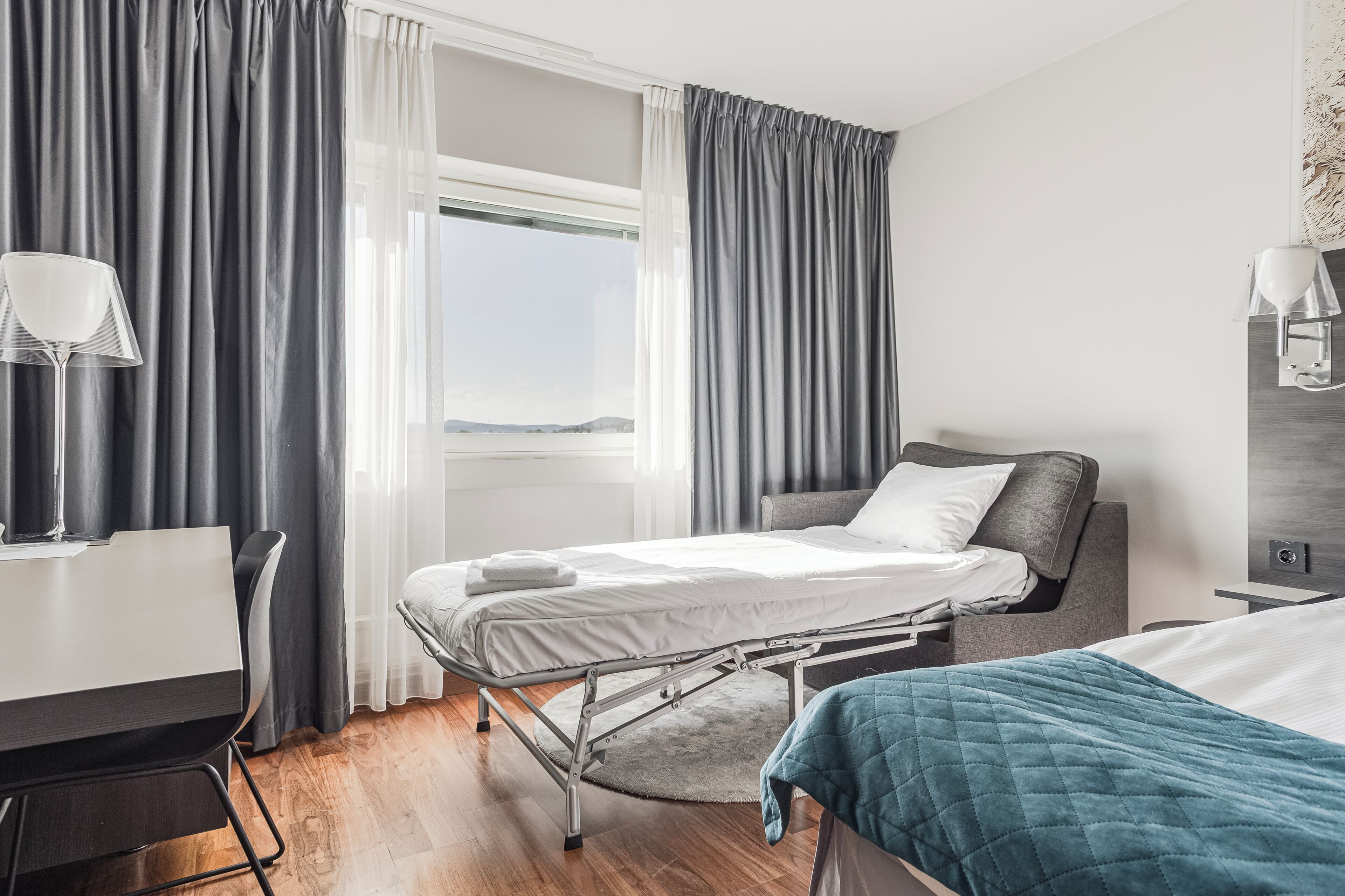 A hotel room with a bed, bedside table, lamp, and a view of the outdoors.
