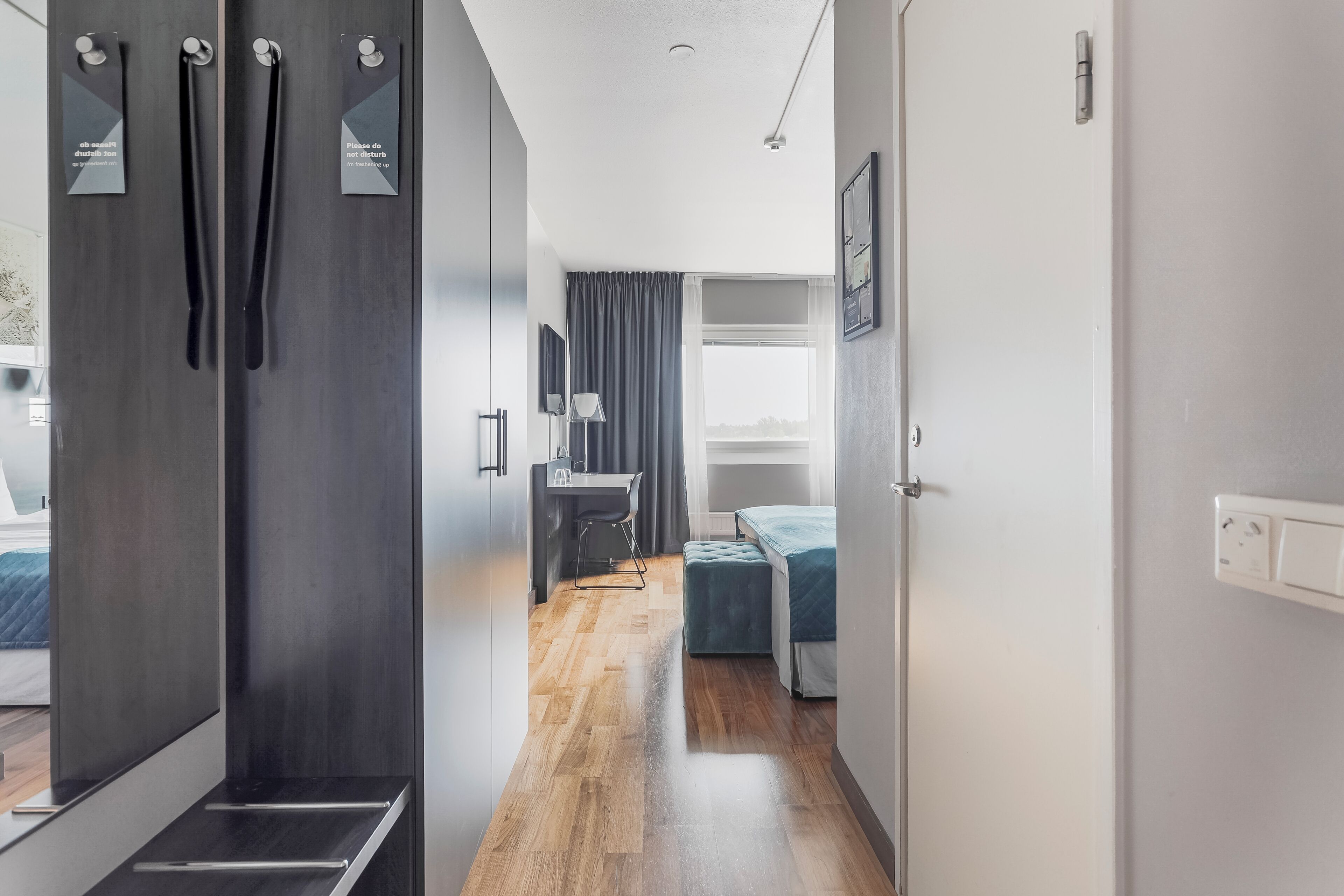 Ein modernes Hotelzimmer mit Holzboden, Schreibtisch mit Stuhl, Bett und Kleiderschrank mit Spiegeln.