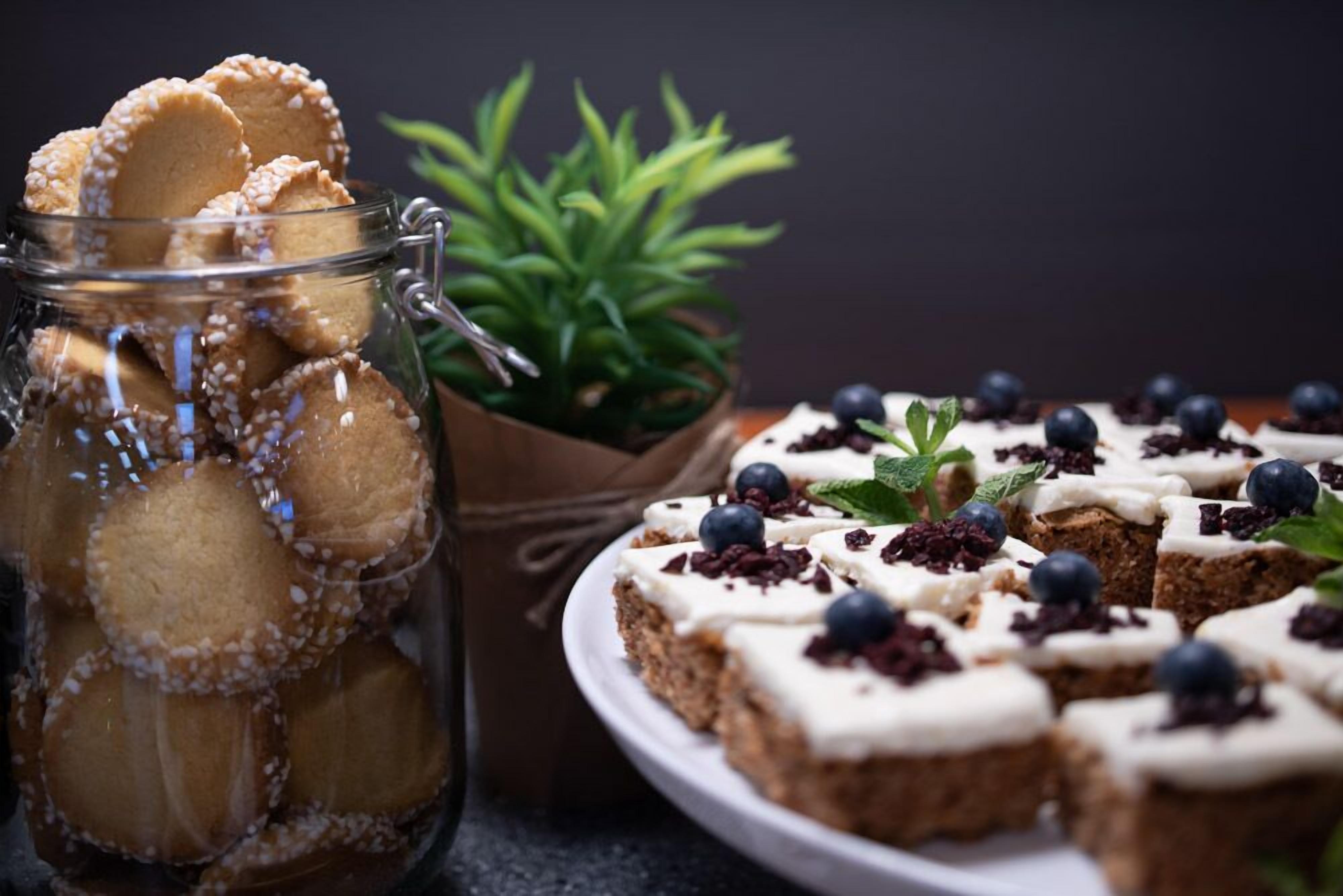 A jar of cookies with white sugar on top, a plate of brownies with blueberries and mint leaves, and a small green plant in the background.