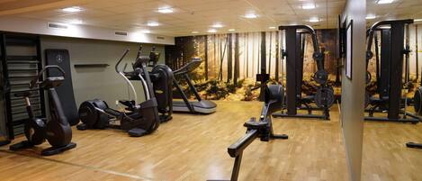 A gym with various exercise equipment, a wooden floor, and a wall with a forest mural.