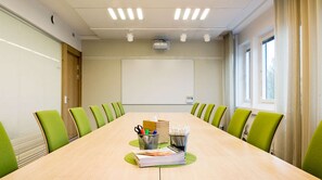 A conference room with a long table, green chairs, a projector screen, and a window.