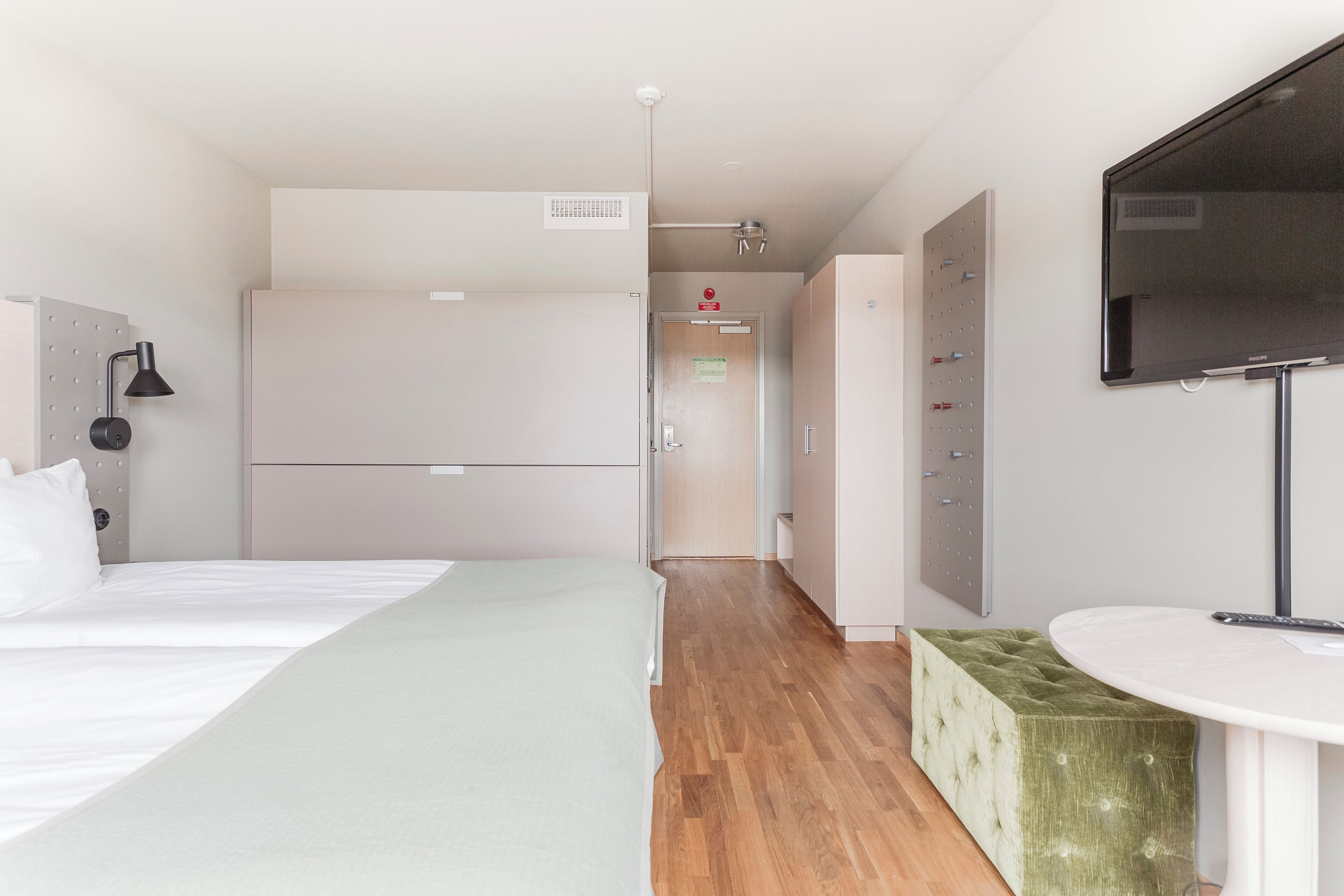 Ett modernt hotellrum med en säng, en grön pall, ett runt bord, en tv och en garderob.
