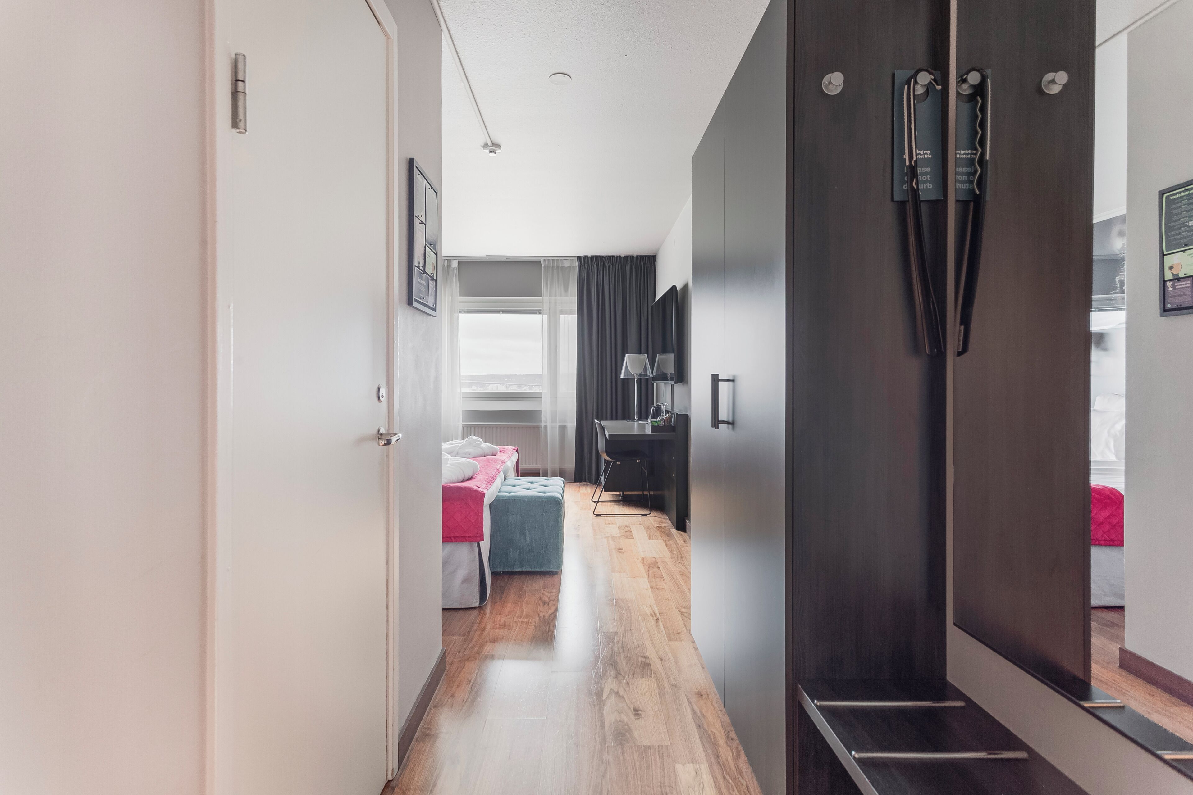 Un couloir d’hôtel moderne avec un plancher en bois, un lit recouvert d’une courtepointe rose et bleue, un bureau avec une lampe et une armoire de couleur sombre.