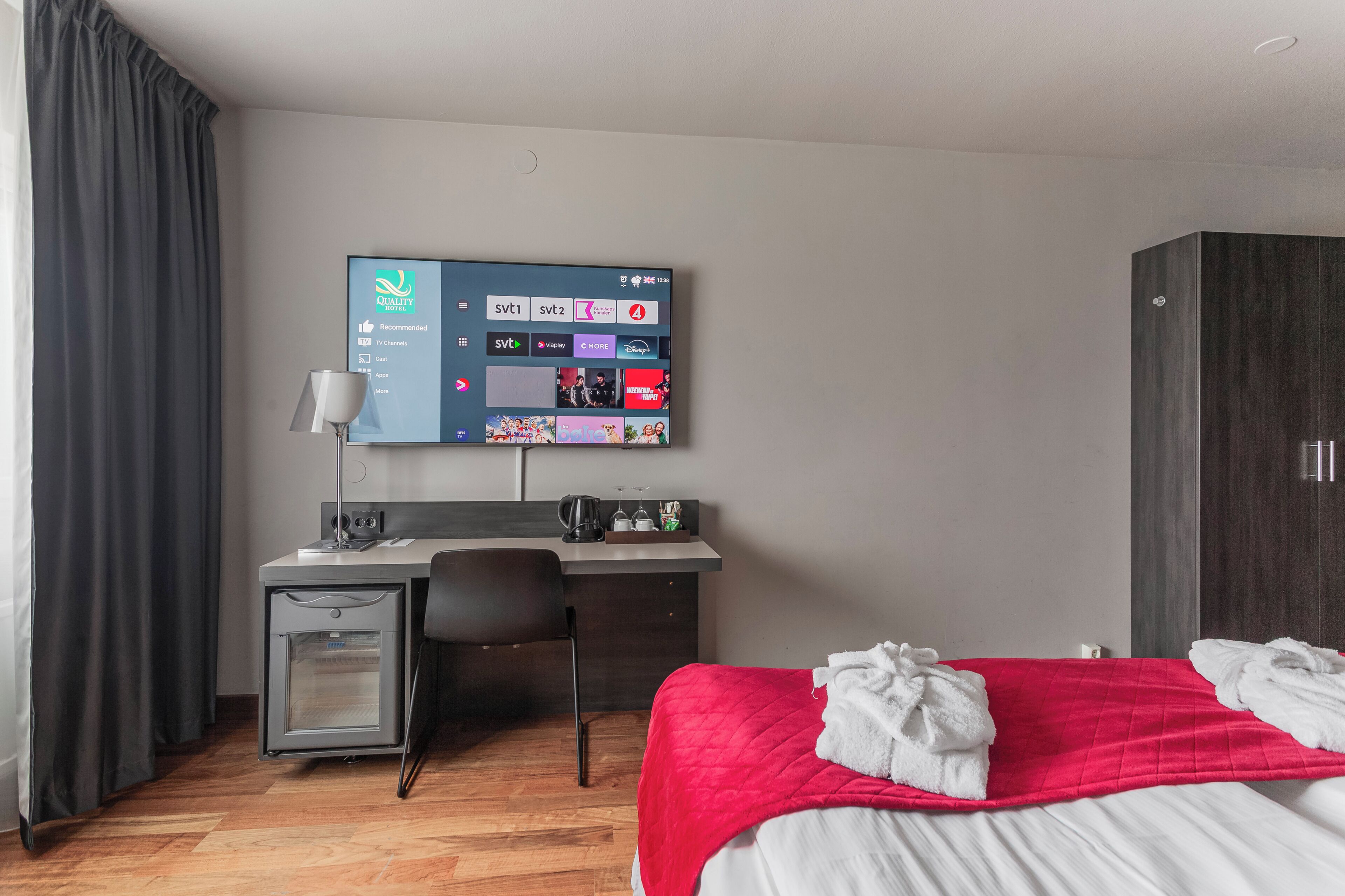 Una habitación de hotel con cama, escritorio, silla, televisor montado en la pared y armario.