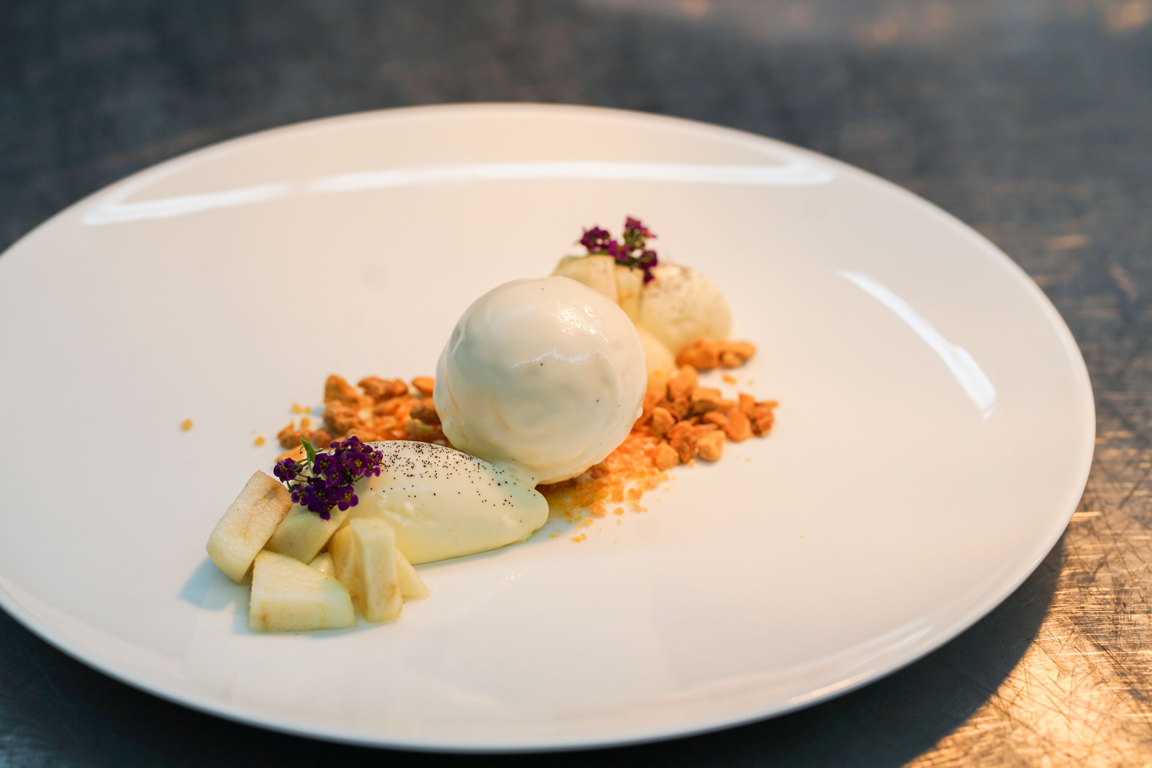 A white plate with a dessert consisting of a scoop of ice cream, diced fruit, and crumbled nuts, garnished with a purple flower.