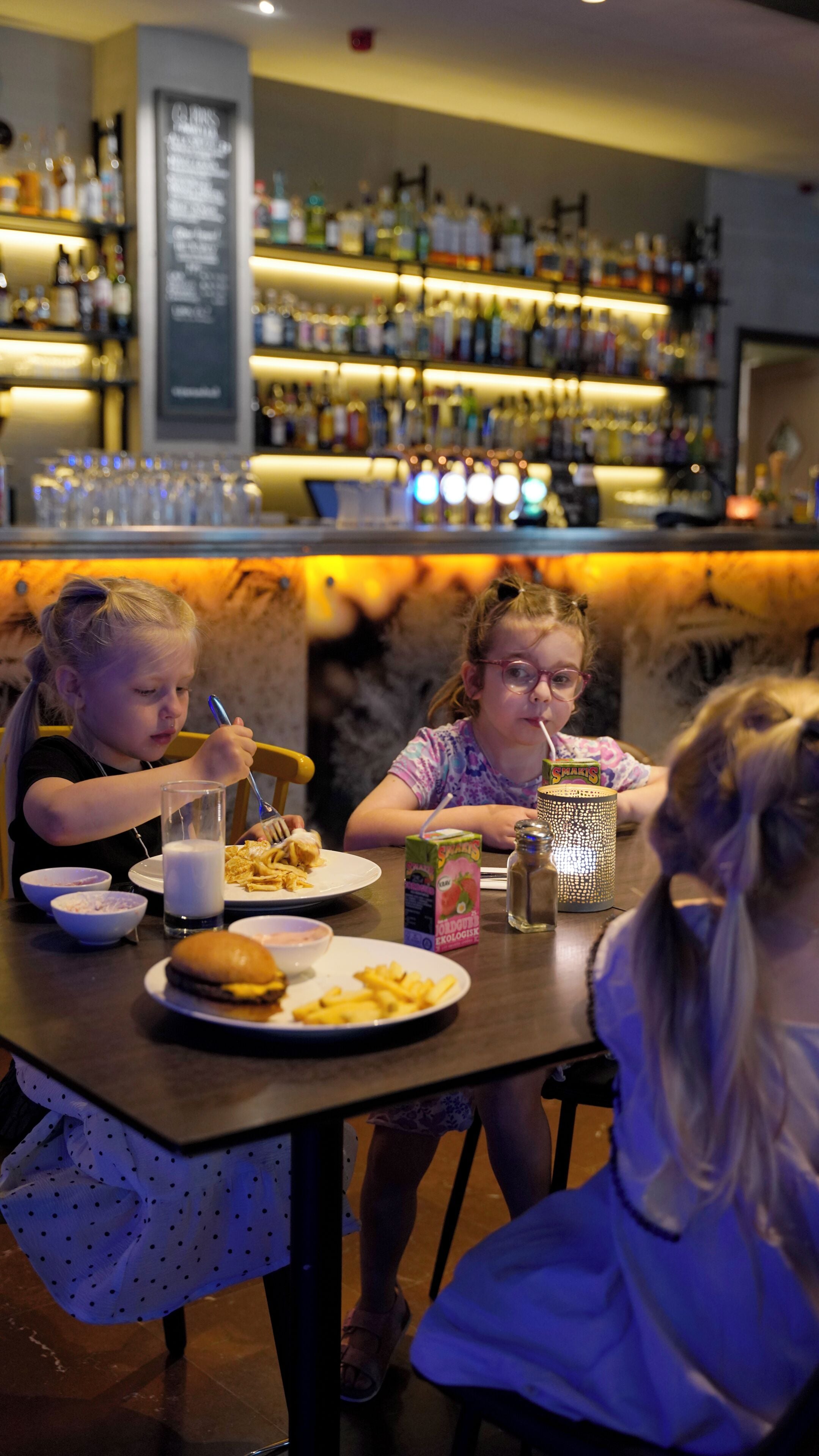 A bar area with shelves of bottles and glasses, a menu board, and a table with food and drinks.