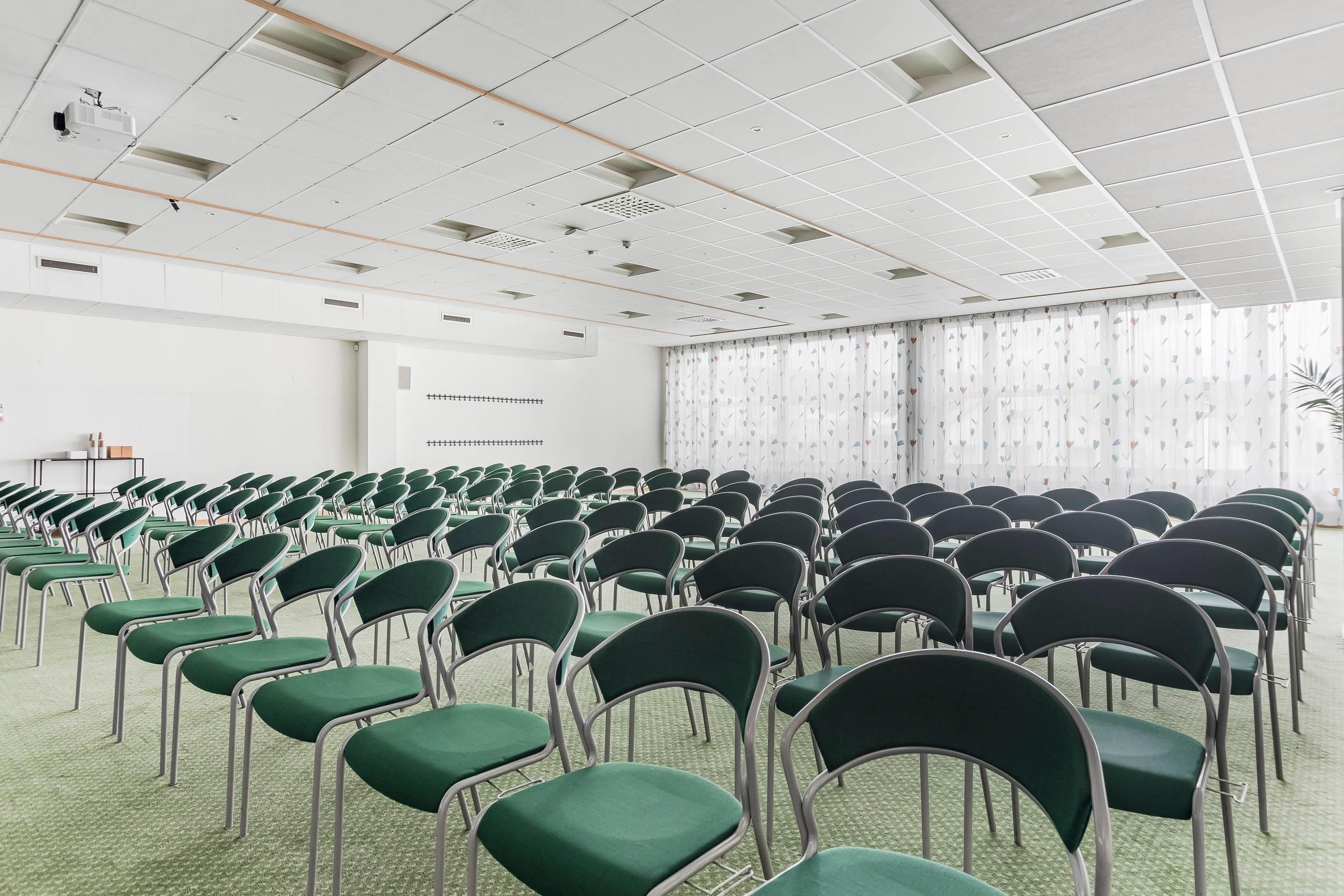 A conference room with rows of green chairs, a white ceiling with grid-like panels, and a large window with patterned curtains.