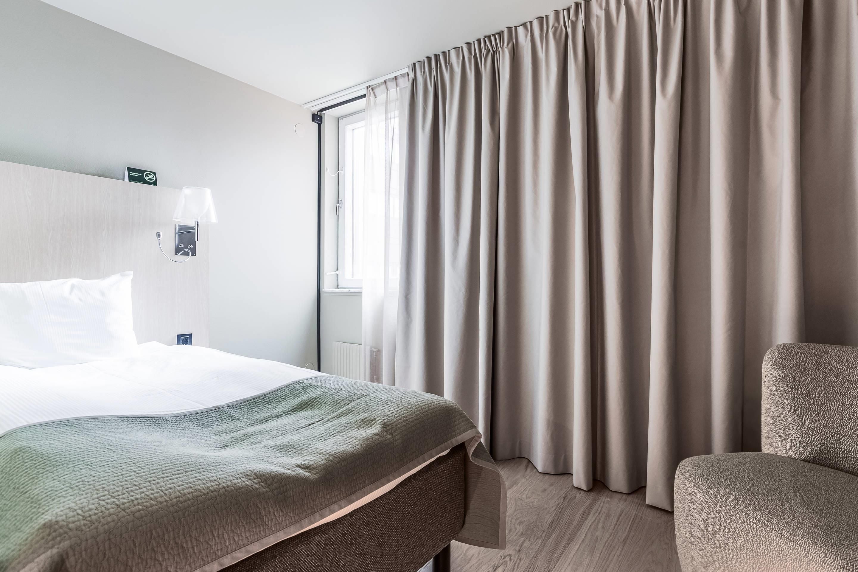 Una habitación de hotel con una cama, una silla, una lámpara y un ventanal con cortinas.
