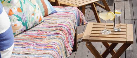 A deck with wooden lounge chairs, a sofa with colorful bedding, and a table with a drink.