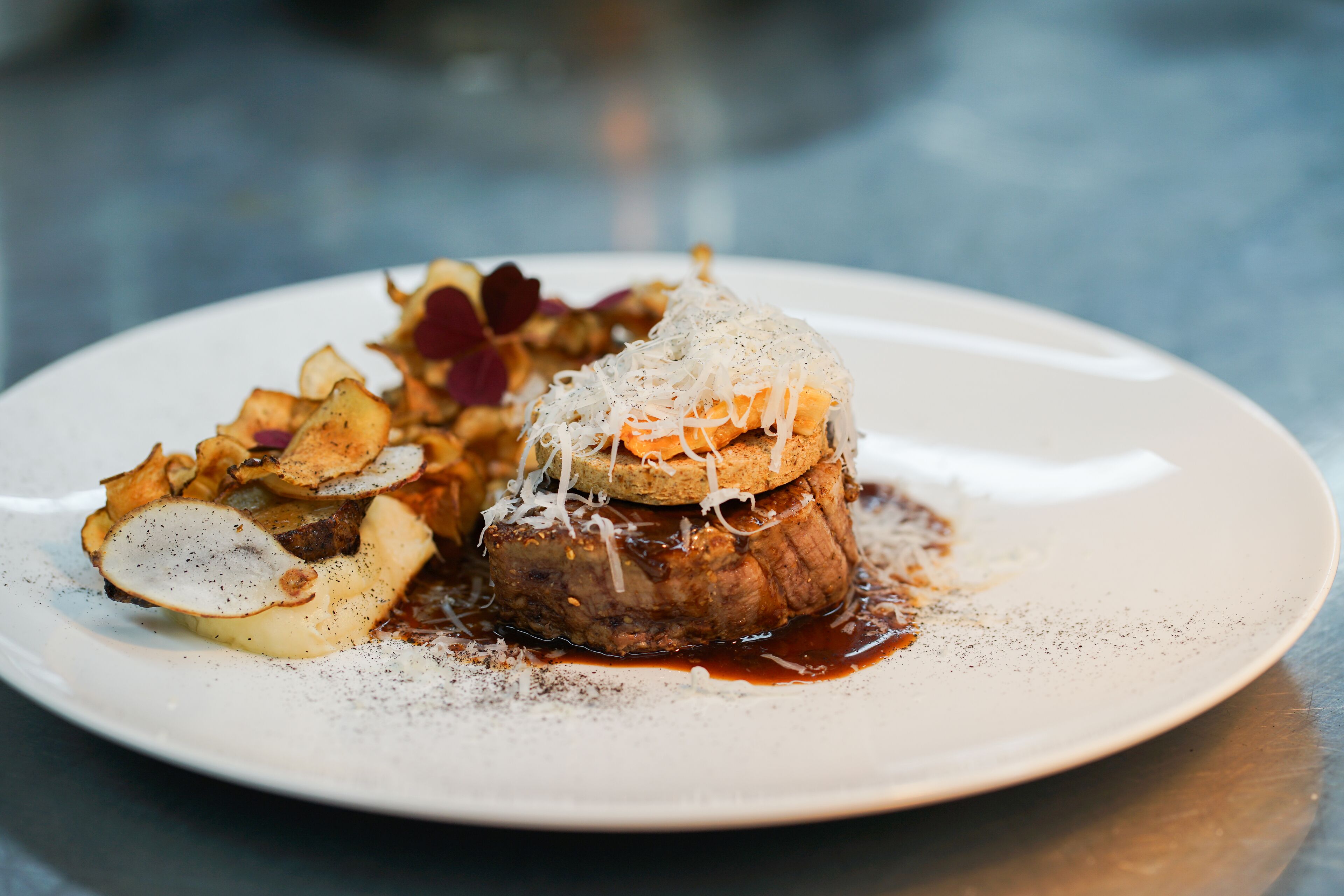A plate of food with a steak topped with cheese and sauce, accompanied by potato slices and a side of greens.