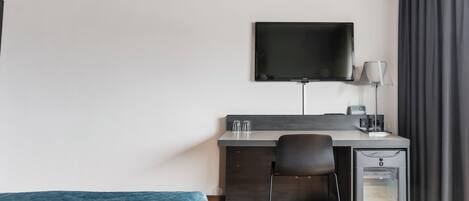 Ett modernt hotellrum med en säng, ett skrivbord med en platt-TV, en stol, en lampa och en minibar.