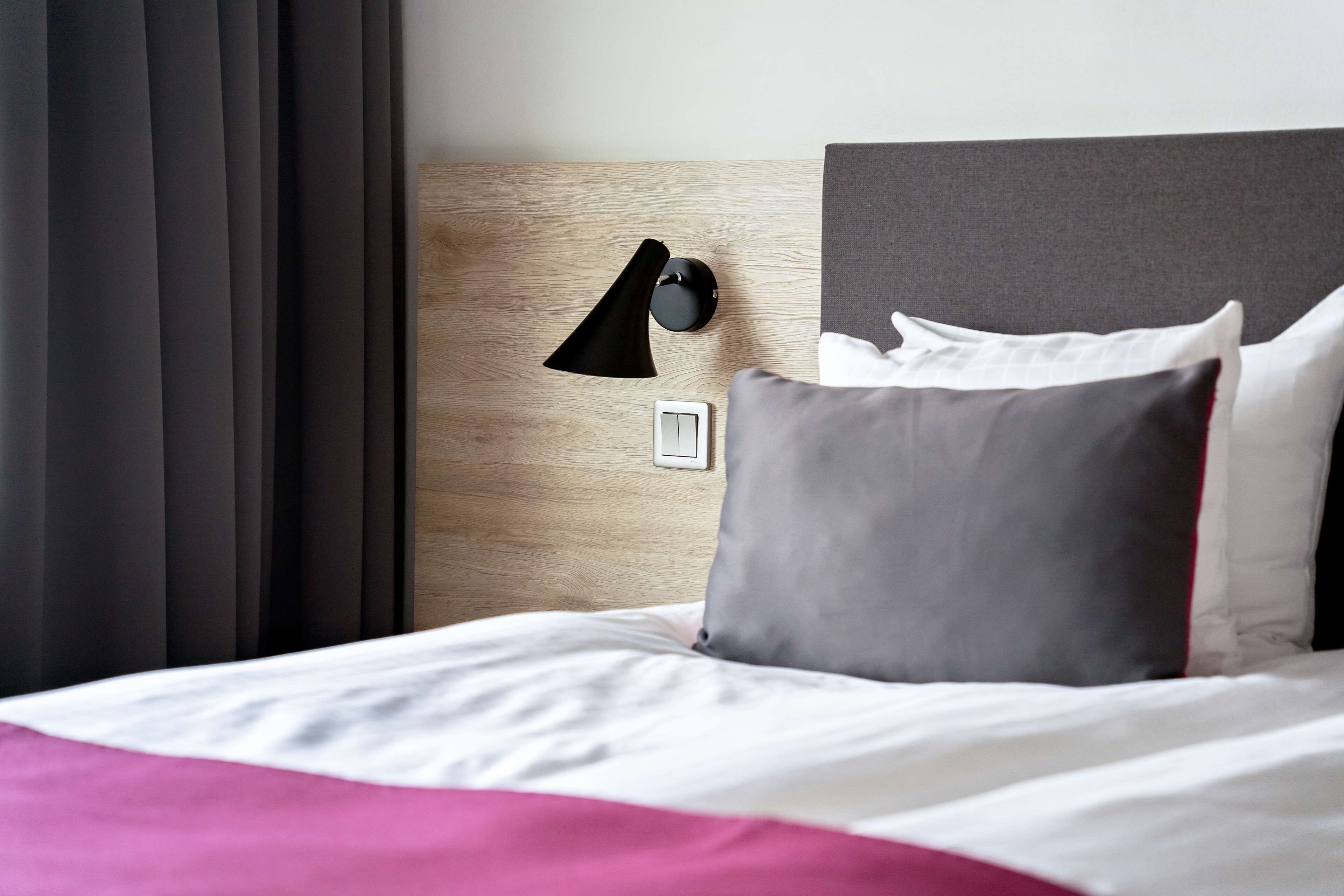 A neatly made bed with white and pink bedding, a gray pillow, and a black wall-mounted lamp.