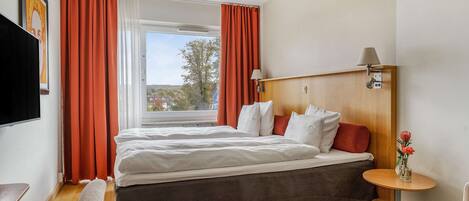 A hotel room with a bed, a bedside table with a lamp, a window with curtains, and a view of the outside.