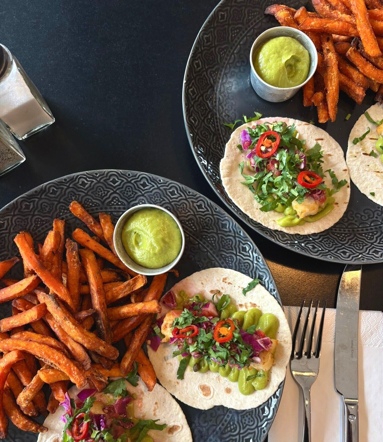 Een tafel met eten, waaronder taco's, friet en groene saus.