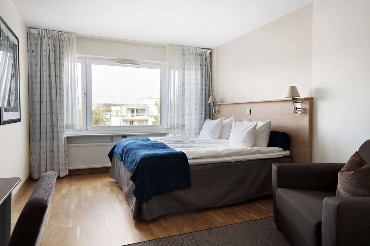 A hotel room with a bed, a desk, a chair, a window with curtains, and a view of buildings outside.