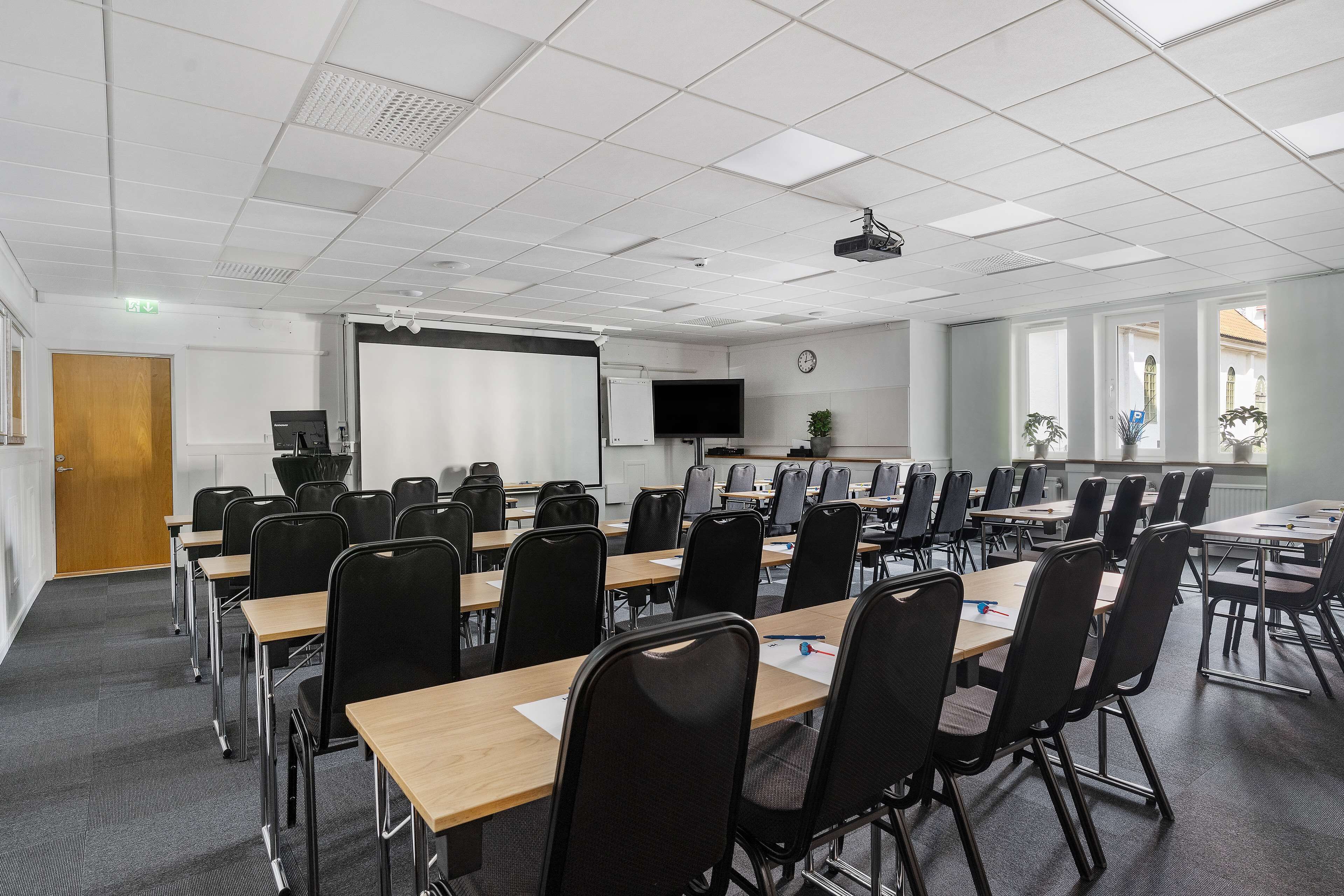 A conference room with multiple tables and chairs, a projector screen, a television, and a clock on the wall.