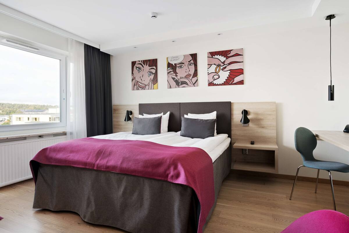 A modern bedroom with a large bed, a desk, and a chair. There are three artworks on the wall, a window with curtains, and a view of the outside.