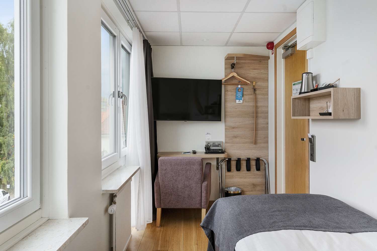 Habitación de hotel pequeña con cama, un escritorio con silla, televisor, estante de madera y ventana con cortinas.