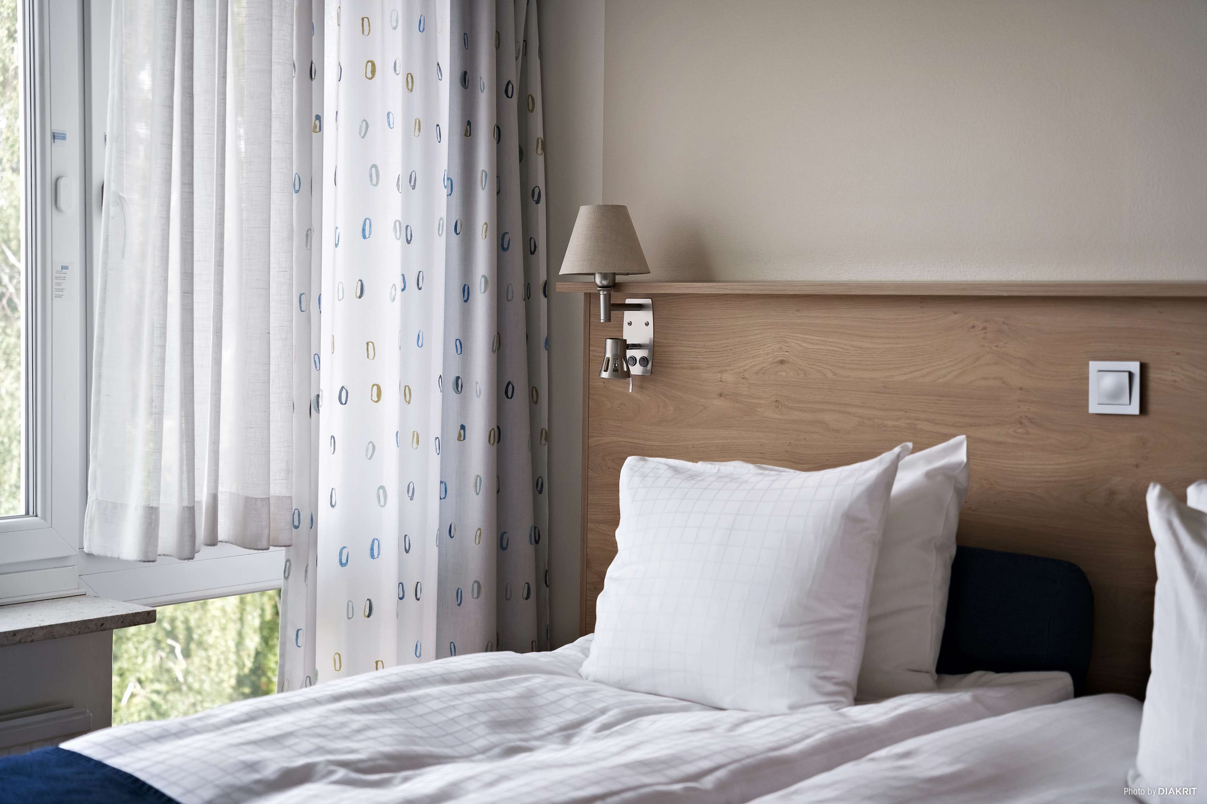 A hotel room with a bed, a window with curtains, a wall-mounted lamp, and a wooden headboard.