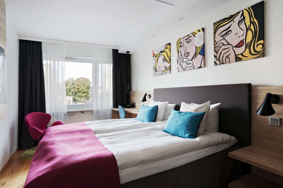 A hotel room with two beds, a desk, a chair, and artwork on the wall.
