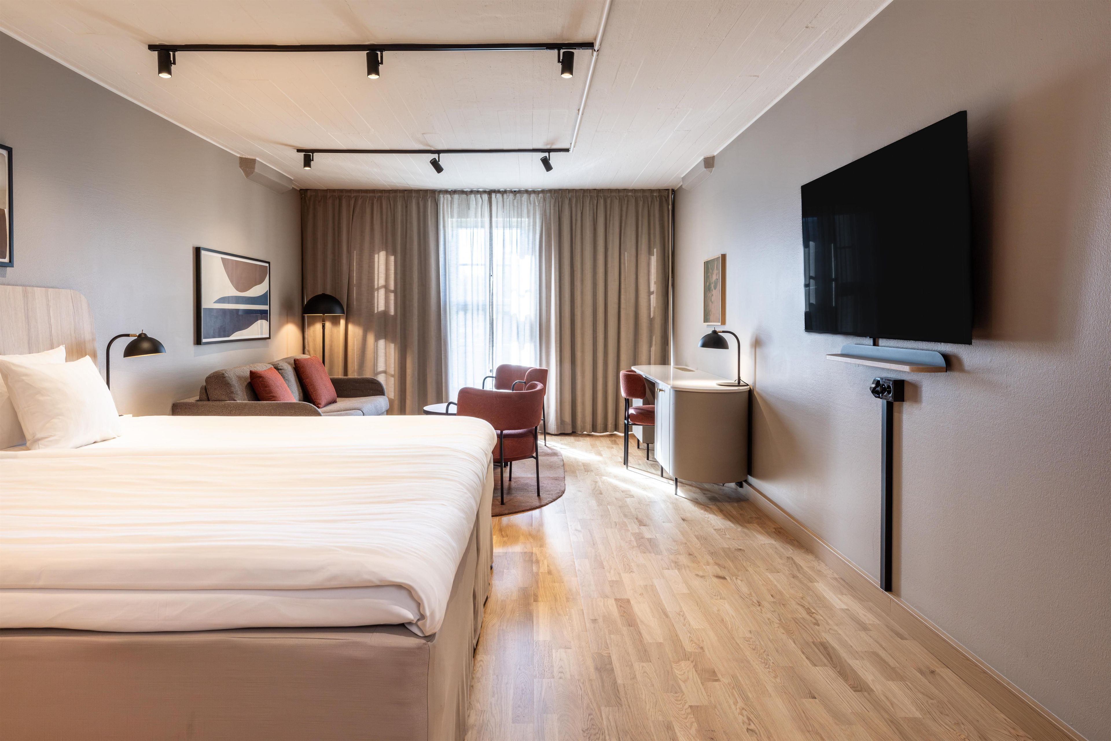Habitación de hotel con cama, escritorio, silla, televisión y ventana con cortinas.