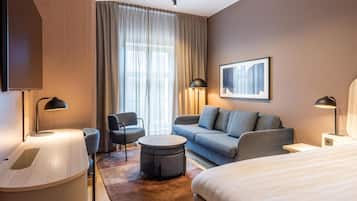 Ett modernt hotellrum med en soffa, två fåtöljer, ett runt soffbord, en säng och ett stort fönster med gardiner.