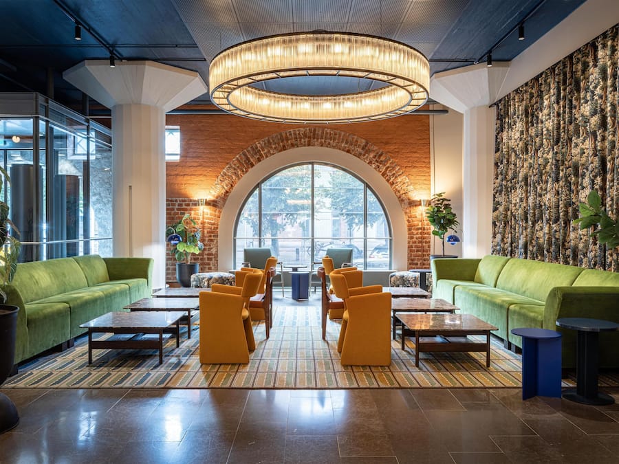 Un hall d’entrée avec un mur en briques, des canapés verts, des fauteuils jaunes et un grand lustre.