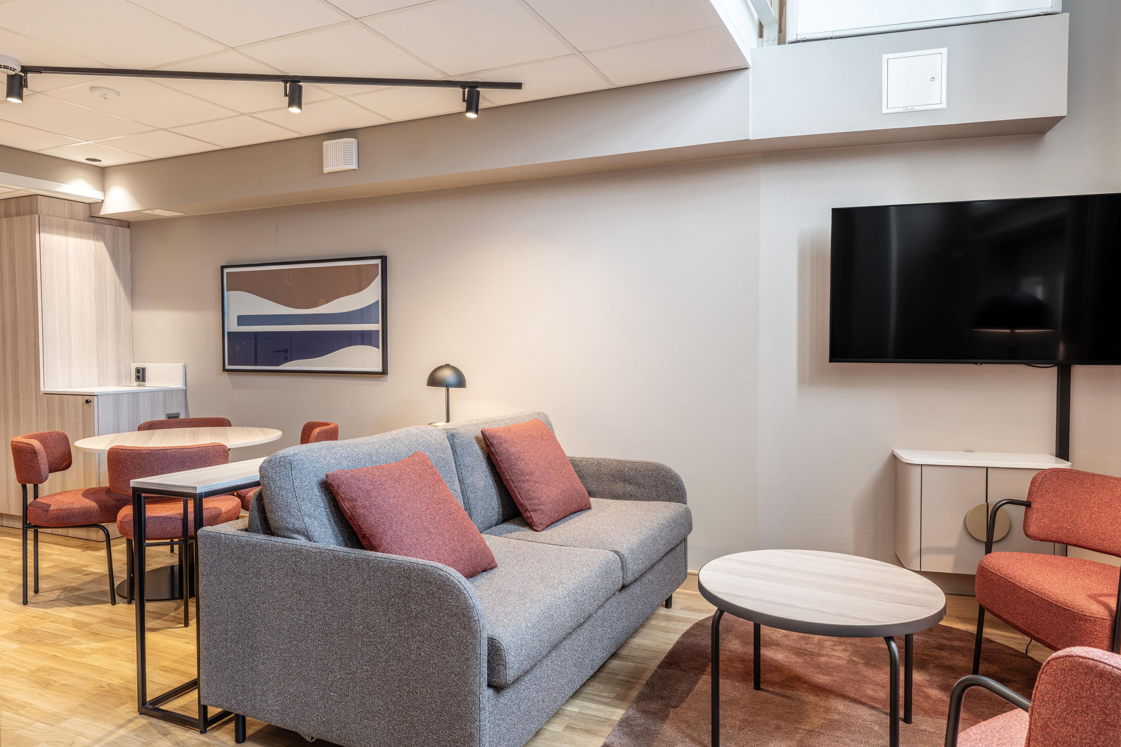 Een moderne hotelkamer met een grijze bank, rode stoelen, een ronde tafel, een flatscreen televisie en een schilderij aan de muur.