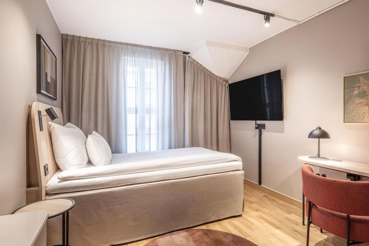 Une chambre d’hôtel moderne équipée d’un lit, d’un bureau, d’une chaise, d’une télévision et d’une fenêtre avec des rideaux.