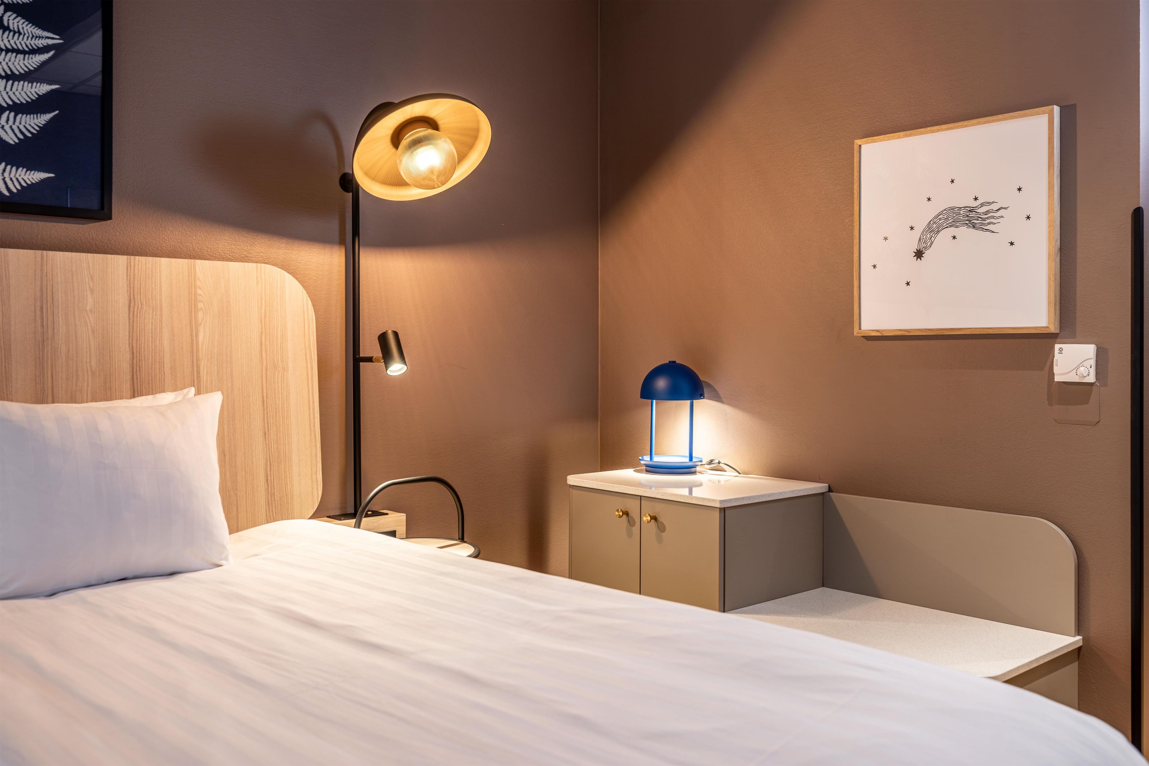 A hotel room with a bed, a desk, a nightstand, a wall-mounted lamp, and a framed artwork.