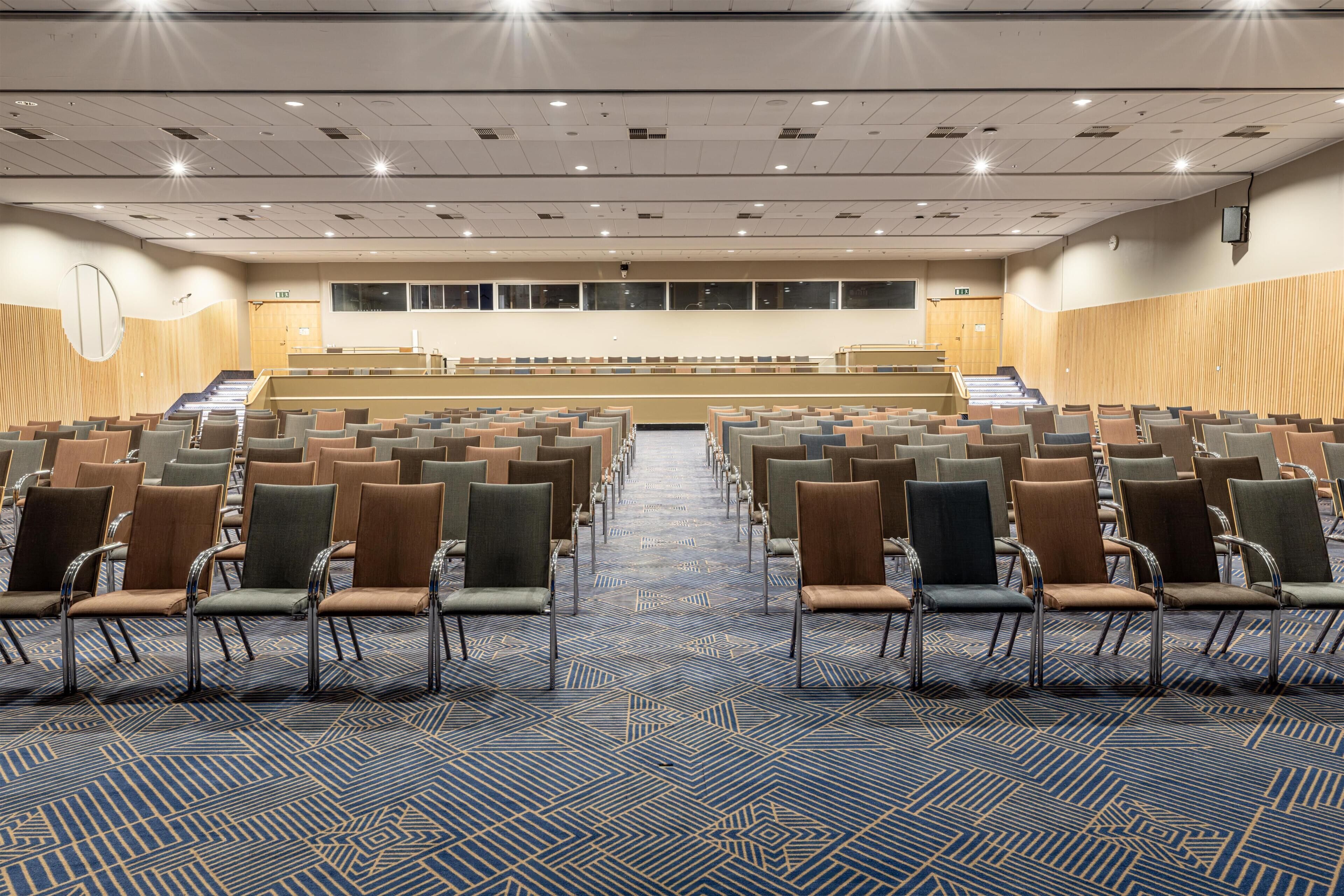 Une salle de conférence avec des rangées de chaises, une scène et un tapis à motifs.