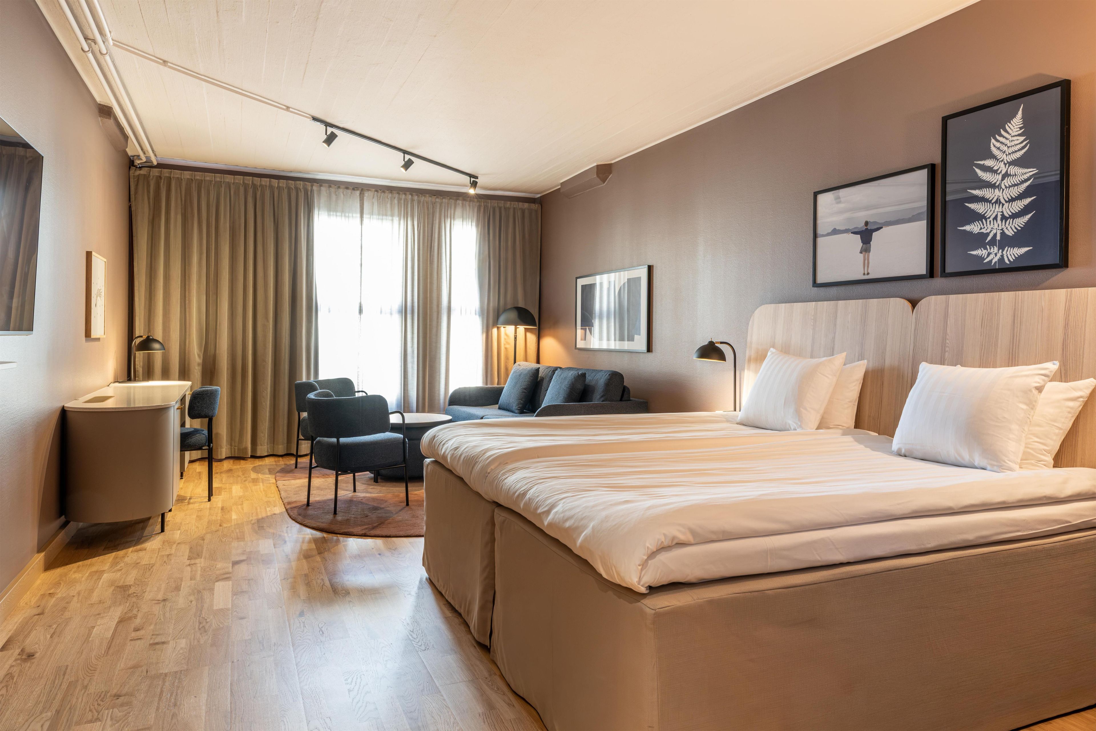 A hotel room with a large bed, a sofa, a desk, and a chair. There are two windows with curtains, a wall-mounted lamp, and framed pictures on the wall.