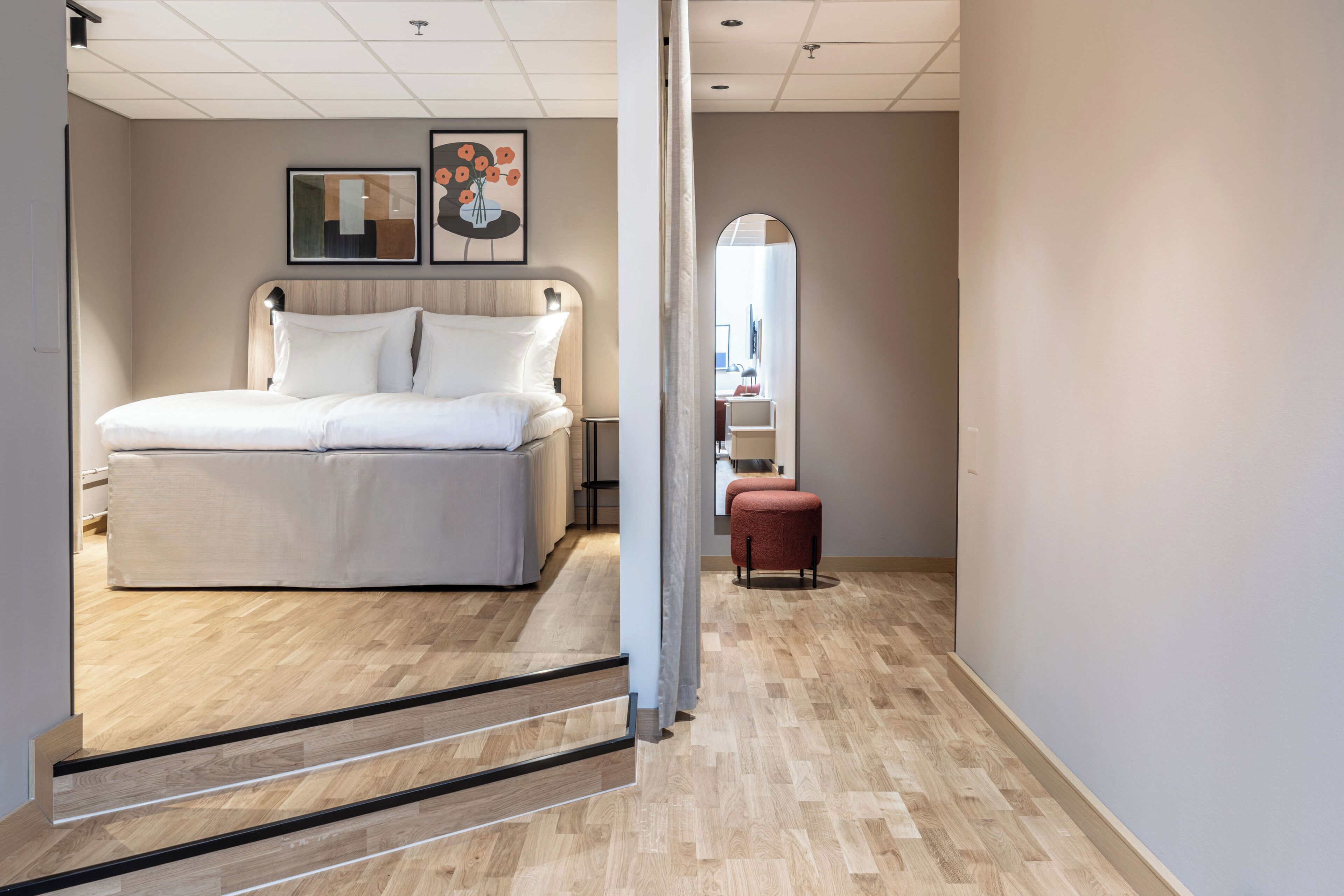 Habitación de hotel con cama, espejo, silla roja y suelo de madera.