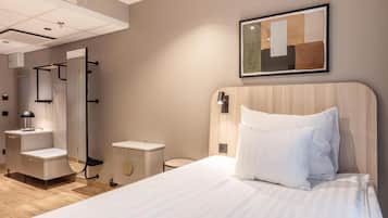 A hotel room with a bed, bedside table, lamp, mirror, and a framed abstract artwork on the wall.