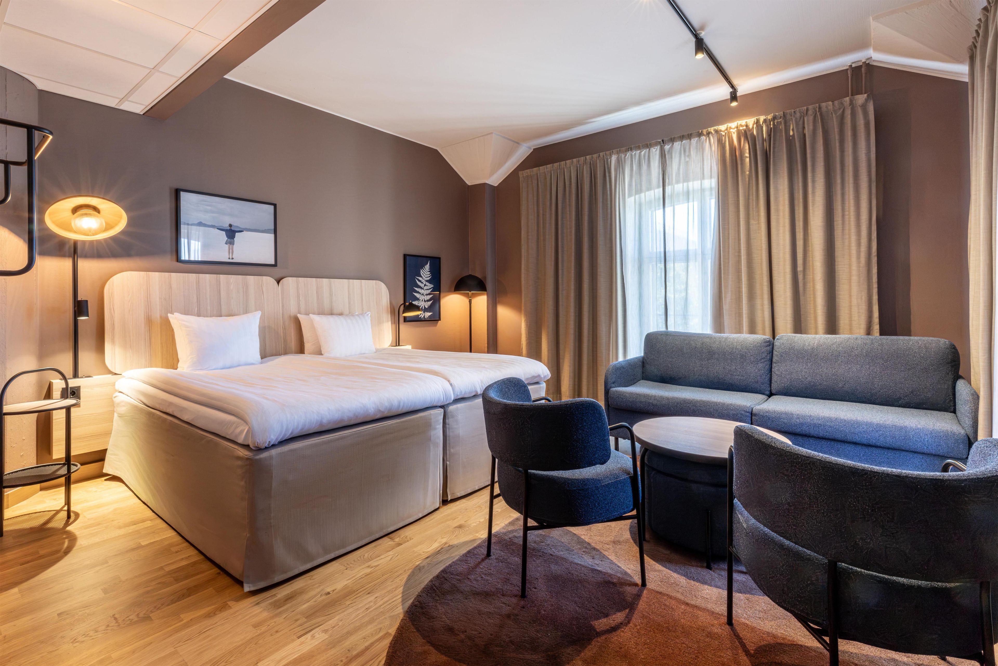 A modern hotel room with a bed, sofa, chairs, and a small table.